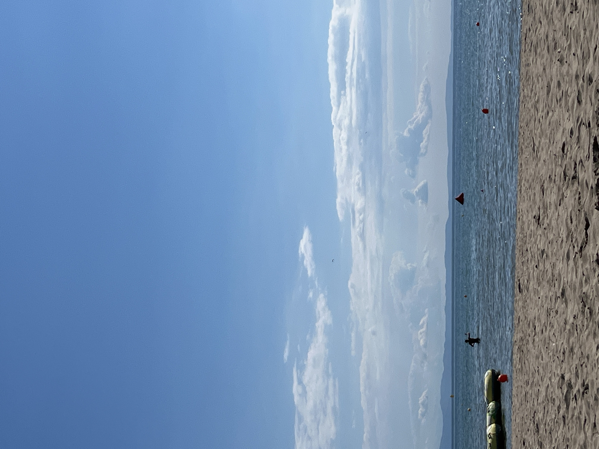 The image depicts a serene beach scene with a clear blue sky dominating the upper portion. Scattered white clouds are visible, with a few birds soaring in the distance. The horizon is marked by a distant mountain range, suggesting an island or coastal location. The sea is calm and blue, with gentle ripples on its surface, reflecting the sky. Several buoys are visible in the water, marking a swimming area or providing navigational guidance. To the left, a person is partially submerged in the water, appearing to be enjoying the sea. In the foreground, a sandy beach stretches across the frame, its texture subtly detailed. The overall atmosphere is one of tranquility and summer relaxation. The location is described as Unknown, Greece.