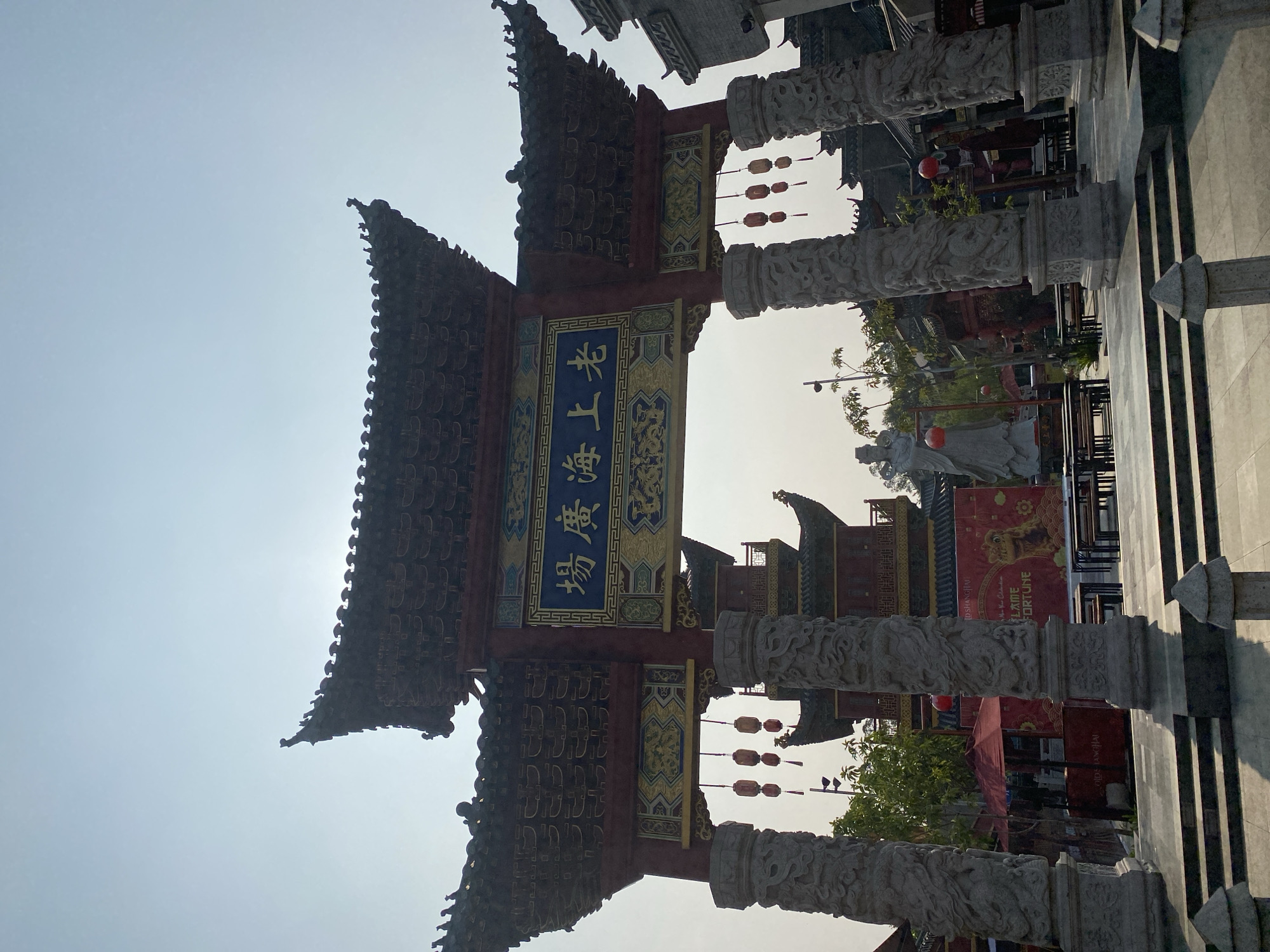 The image depicts an ornate Chinese-style archway with traditional tiled roofs and intricate carvings on its supporting pillars. The archway features a blue sign with yellow Chinese characters. Red lanterns hang from the archway, and a tiered pagoda-like structure is visible behind it. In the background, there are several buildings with similar architectural elements, suggesting an East Asian-themed area. In front of the pagoda, a large banner with Chinese characters and an image of a mythical creature is displayed, along with tables and chairs, indicating a potential dining or performance space. A statue of a person in traditional attire stands to the left of the banner. The overall setting suggests a cultural or entertainment district. The location is identified as the "Hoo Keng" or "Old Shanghai" area in North Jakarta, Indonesia.