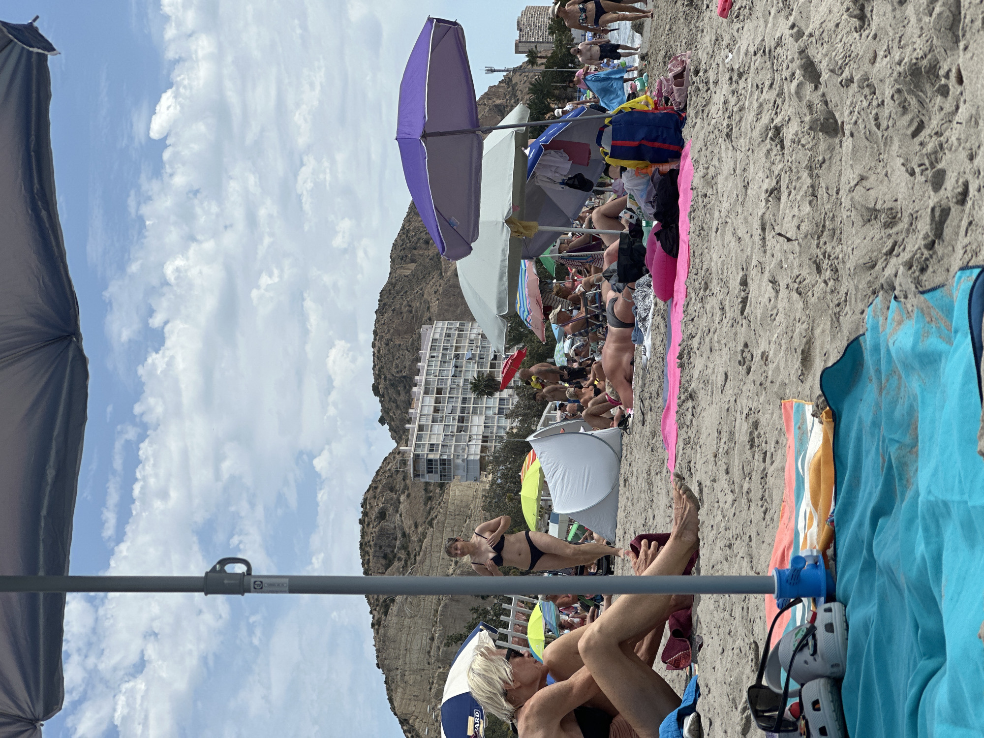 The image depicts a crowded beach scene under a partly cloudy sky. In the foreground, a blue towel is laid out on the sand, with a pair of white Crocs and sunglasses placed on top. To the left, a woman with blonde hair and sunglasses is sitting on the sand, facing away from the camera, wearing a black bikini top and part of a swimsuit bottom. Behind her, another woman in a black bikini is walking, with a small white tent set up on the sand nearby. In the background, several beach umbrellas are visible, as well as a number of people relaxing on the beach. A large, white building with multiple balconies stands against a rocky hillside. The overall scene is one of a busy beach resort on a sunny day.