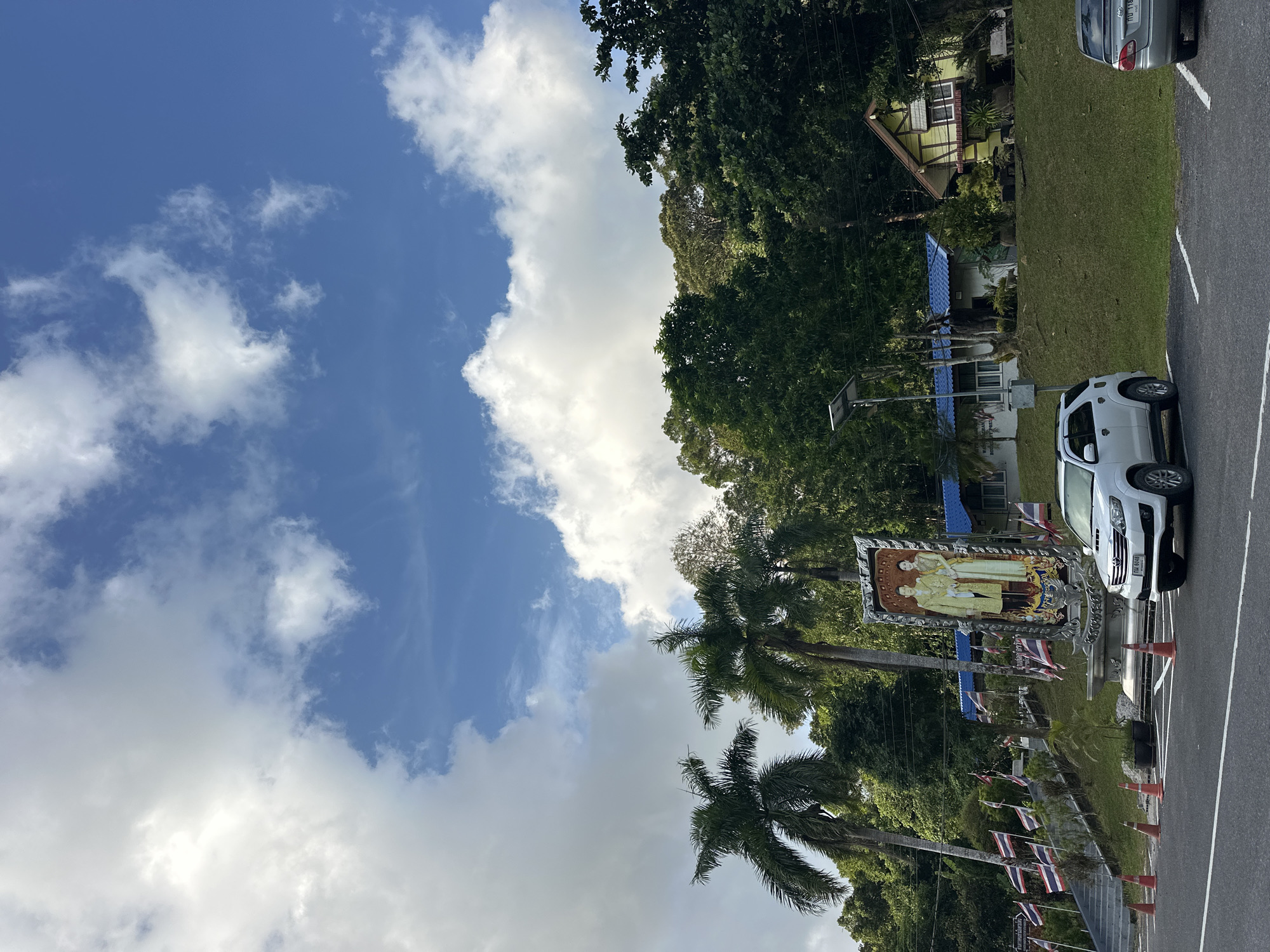 The image depicts a scene in Tambon Wang Prachan, Thailand, under a bright blue sky with scattered white clouds. In the foreground, a paved road with white parking lines is visible, with a white SUV parked in the right lane and a silver sedan parked to its right. Orange traffic cones are placed along the edge of the road.
To the left of the road, a sloped grassy area leads up to a pathway lined with Thai flags. Further up, a large framed portrait of a man and a woman in regal attire stands prominently. Behind the portrait, there are palm trees and lush green foliage. A low-rise building with a blue roof is visible behind the trees, and a quaint yellow house with a distinctive roofline is situated on the right side of the frame, partially obscured by trees.
The overall impression is of a scenic, possibly historical or governmental site, with elements of local culture and natural beauty.