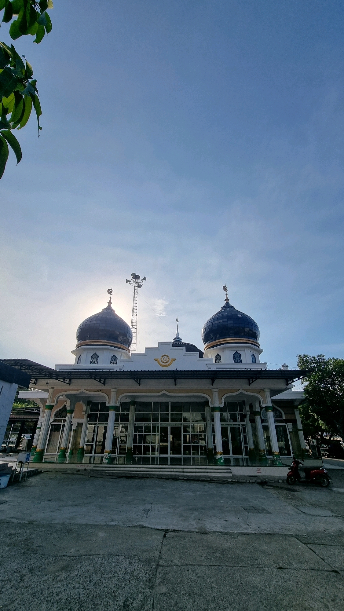 A wide-angle, low-angle shot captures a mosque in Blangrambong, Indonesia, under a clear blue sky. The mosque features two prominent dark blue domes topped with crescent moons. The facade is white with ornate columns and large glass windows. A tall tower with loudspeakers stands between the domes, with the sun partially obscuring it. To the left, green leaves hang from a branch, and to the right, a red motorcycle is parked near a tree. The ground in front of the mosque is paved with concrete. The image appears to have been taken in the morning or late afternoon due to the sun's position.