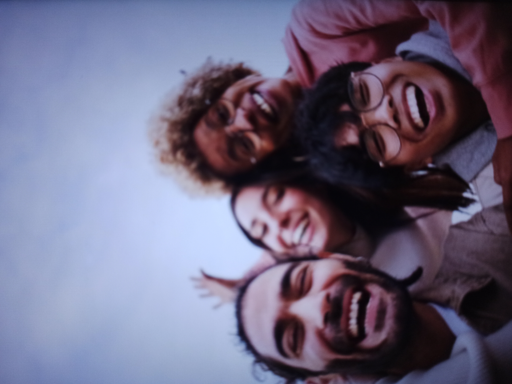 A low-angle, close-up shot captures four young adults posing for a selfie against a pale blue sky. The individuals are grouped closely, with two in the foreground and two behind them.
In the foreground, a man with dark, curly hair and a beard smiles widely, his mouth open in a laugh. He is wearing a light-colored scarf or cowl neck and a brown jacket. To his right, a woman with long, dark hair is also laughing, her eyes closed and head tilted back. She is wearing a light-colored top.
Behind them, a woman with curly blonde hair and glasses laughs heartily, her head tilted. She is wearing a pink hoodie. To her right, a person with short, dark hair and glasses is laughing, their mouth open. They are wearing a gray hoodie. The sky is the dominant background element, appearing overcast or hazy. The overall impression is one of joy and camaraderie.