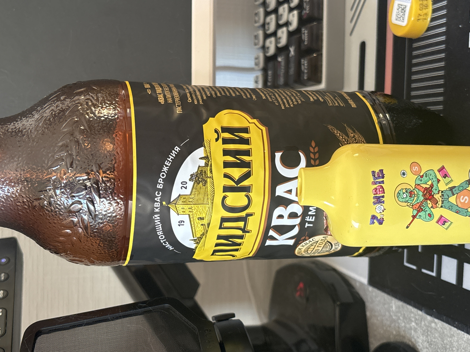 A brown glass bottle labeled "Lidskiy Kvas" sits horizontally on a desk next to a yellow bottle with a cartoon zombie on it. The Lidskiy Kvas bottle has a black label with yellow writing in Russian. In the background, there is a black keyboard and a gray and white object. The photo does not provide any information to indicate that it was taken in the city of Vsevolozhsk, Russia.