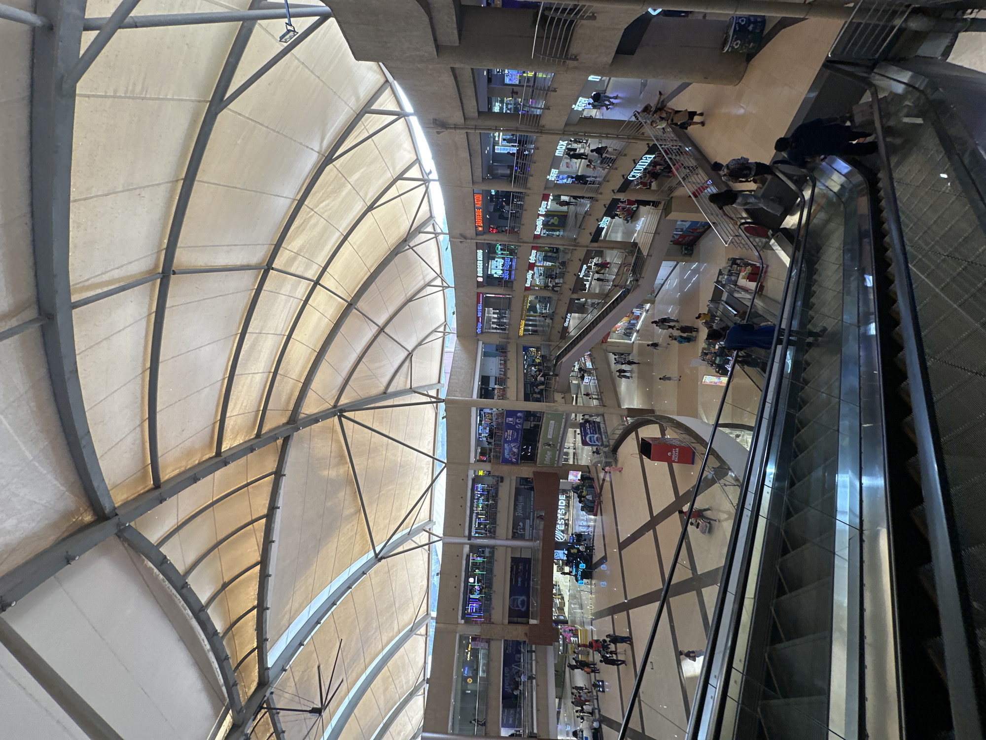 The image depicts the interior of a shopping mall located in Kalyan-Dombivli, India. The mall has multiple floors, visible with various retail outlets and shops. People are seen walking and using escalators. The mall has a large, arched roof structure made of metal supports and a fabric or membrane-like material. Escalators are positioned on the right side of the image. The lighting seems to be a combination of natural light coming through the roof and artificial lighting within the mall.