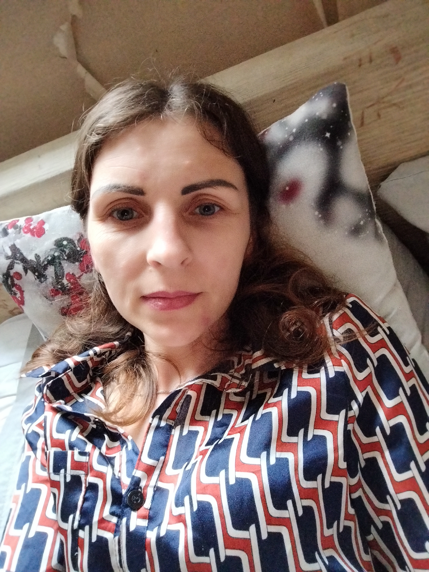 The image is a close-up, selfie-style photograph of a woman lying down, with her head resting on two pillows. The woman has medium-length, wavy brown hair, and is looking directly at the camera. She is wearing a patterned shirt with blue, red, and white geometric designs. One pillow to her left has a floral pattern with red and white flowers on a white background. The pillow to her right is partially visible and appears to have a celestial pattern with a face-like image. The background consists of a textured ceiling and wooden beams, suggesting an indoor setting, possibly a bedroom. No specific location in Vinnytsia, Ukraine, can be determined from the visual information.