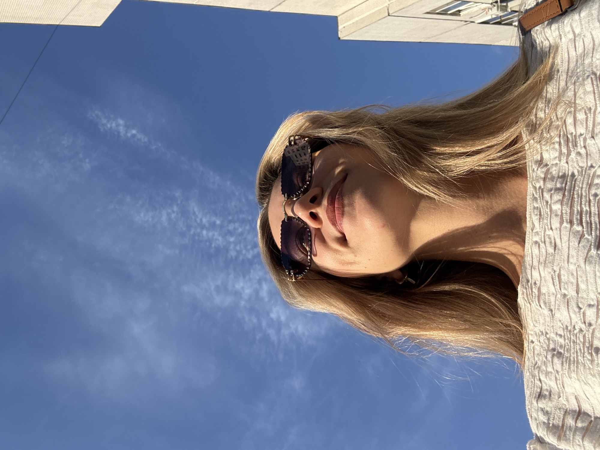 The image depicts a woman with blonde hair, wearing sunglasses and a light-colored, possibly white, textured top. She is positioned outdoors with a clear blue sky overhead, punctuated by a few wispy clouds. The perspective is from below, looking upwards at the woman. Part of a building structure, possibly made of concrete or stone, is visible in the upper left corner of the frame. The location is in an urban setting, indicated by the building, but the city is unknown.