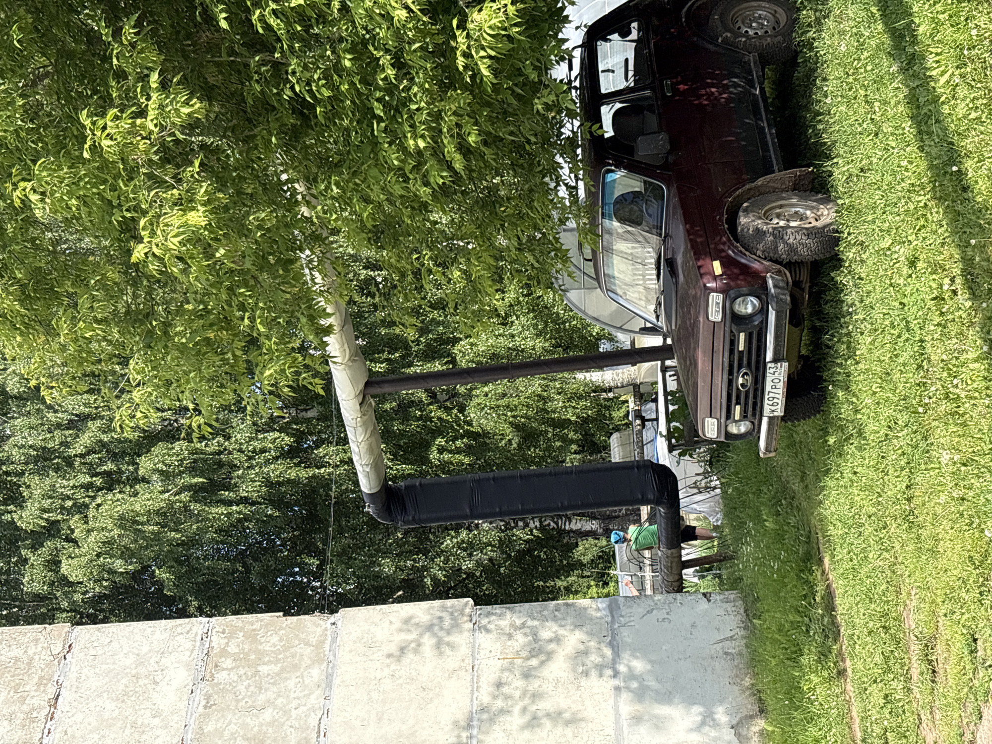 In the village of Kumyon, Russia, a dark red Lada Niva is parked on a grassy area next to a concrete building and under the shade of a large tree. The car's license plate is visible and reads "K697PO43". A person wearing a green shirt and a blue head covering is standing in the background, near some pipes. Overhead, large pipes, insulated with white material and one section wrapped in black material, are supported by a metal frame. The environment appears to be a residential or utility area with lush green grass and dense foliage from the trees.