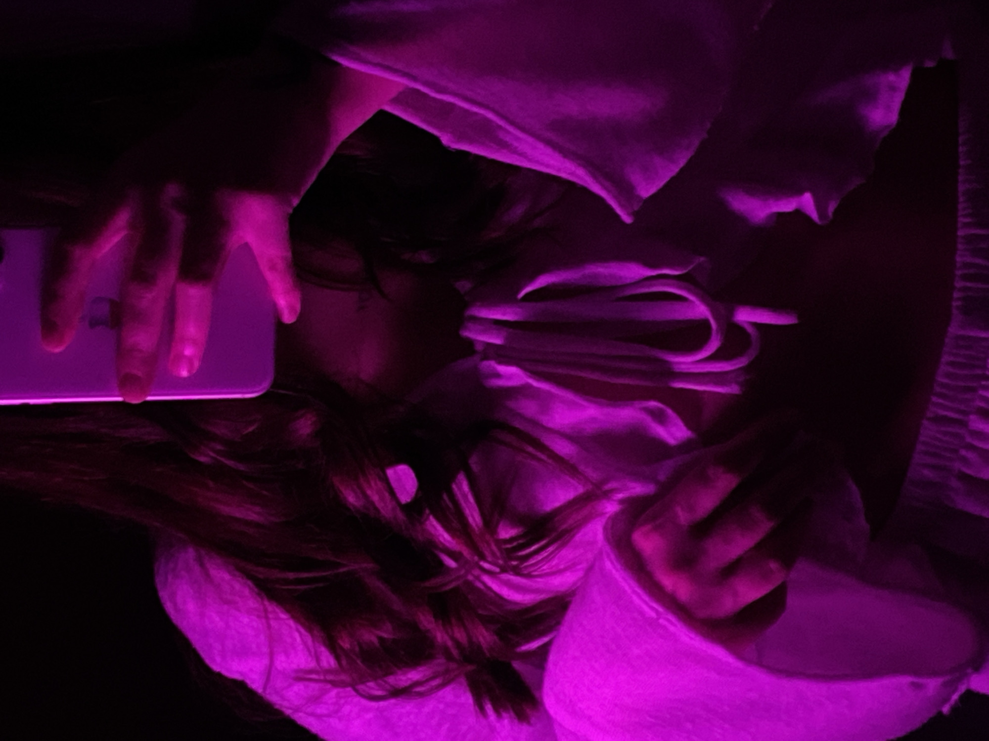 A person is lying down and looking at a white smartphone in their left hand. The person is wearing a white shirt or blouse that is tied in the middle. The scene is lit with a purple light. The location is not specified in the image and cannot be determined.