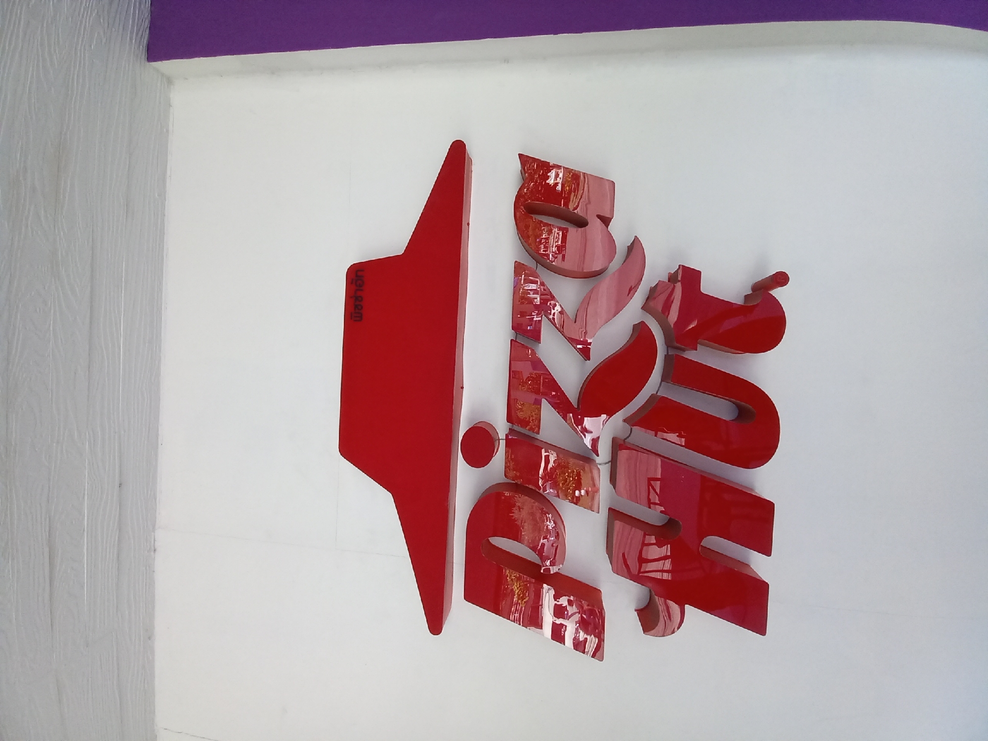 This is a low-angle, eye-level, outdoor, medium shot of a red "Pizza Hut" sign mounted on a white wall. The sign features the iconic red Pizza Hut roof shape above the red, three-dimensional letters spelling "PIZZA HUT." The letters are shiny and reflect the surrounding environment, which appears to be a street with some vehicles. There is also some Thai text on the red roof shape. The wall is flat and white, with a light gray, textured ceiling or upper wall section above it. To the right of the white wall is a purple vertical panel. The lighting suggests it is daytime, with some shadows on the wall and the sign. The overall scene appears to be in front of a Pizza Hut restaurant in Baan Bo Phut, Thailand.