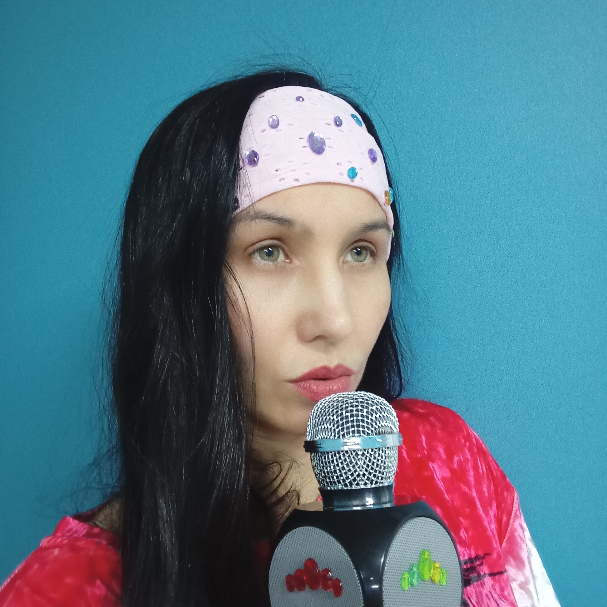 An indoor, eye-level shot captures a woman holding a microphone against a plain, vibrant teal-blue wall. The woman has long, dark hair, light-colored eyes (appearing greenish-hazel), and is wearing red lipstick. She has a serious, contemplative expression, looking off to the right side of the frame. A pink headband adorned with various colored, reflective jewels (purple, blue, yellow) is visible on her forehead, pushing back some of her hair. She is wearing a red top, which appears to be made of a soft, textured fabric like velvet. In front of her, she holds what appears to be a karaoke or toy microphone. It features a silver mesh top, a light blue band around its mid-section, and a black base with speaker grilles. The speaker grilles are decorated with colorful, raised plastic elements, specifically red on the left grille and green on the right. There is no visible text or specific location cues within the image.