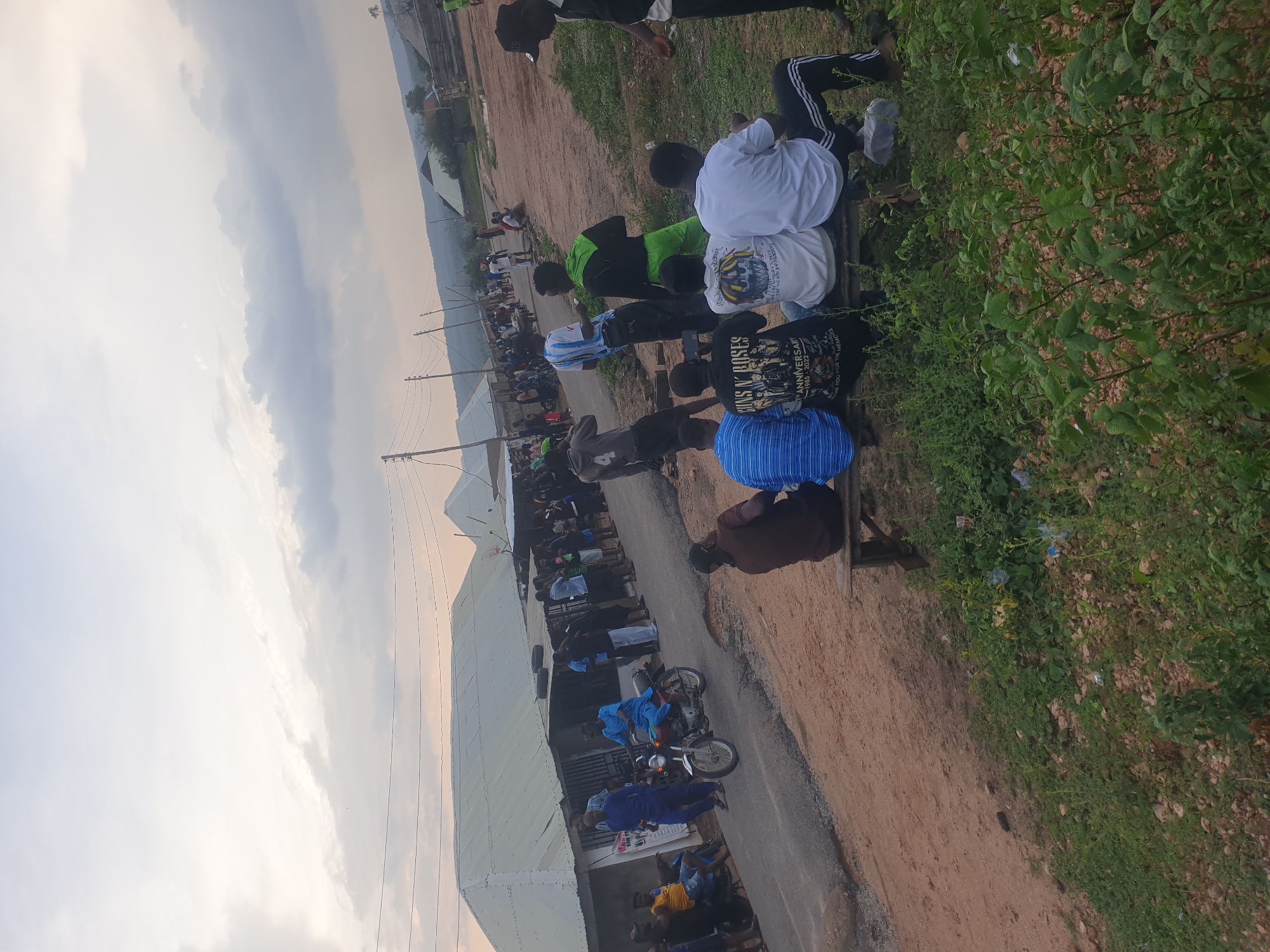 The image shows a crowded outdoor scene in Zango-ndaji, Nigeria. Many people are gathered along a road, some sitting on benches near green vegetation. Other people are walking or standing near buildings. A few motorcycles are present. The sky is cloudy.