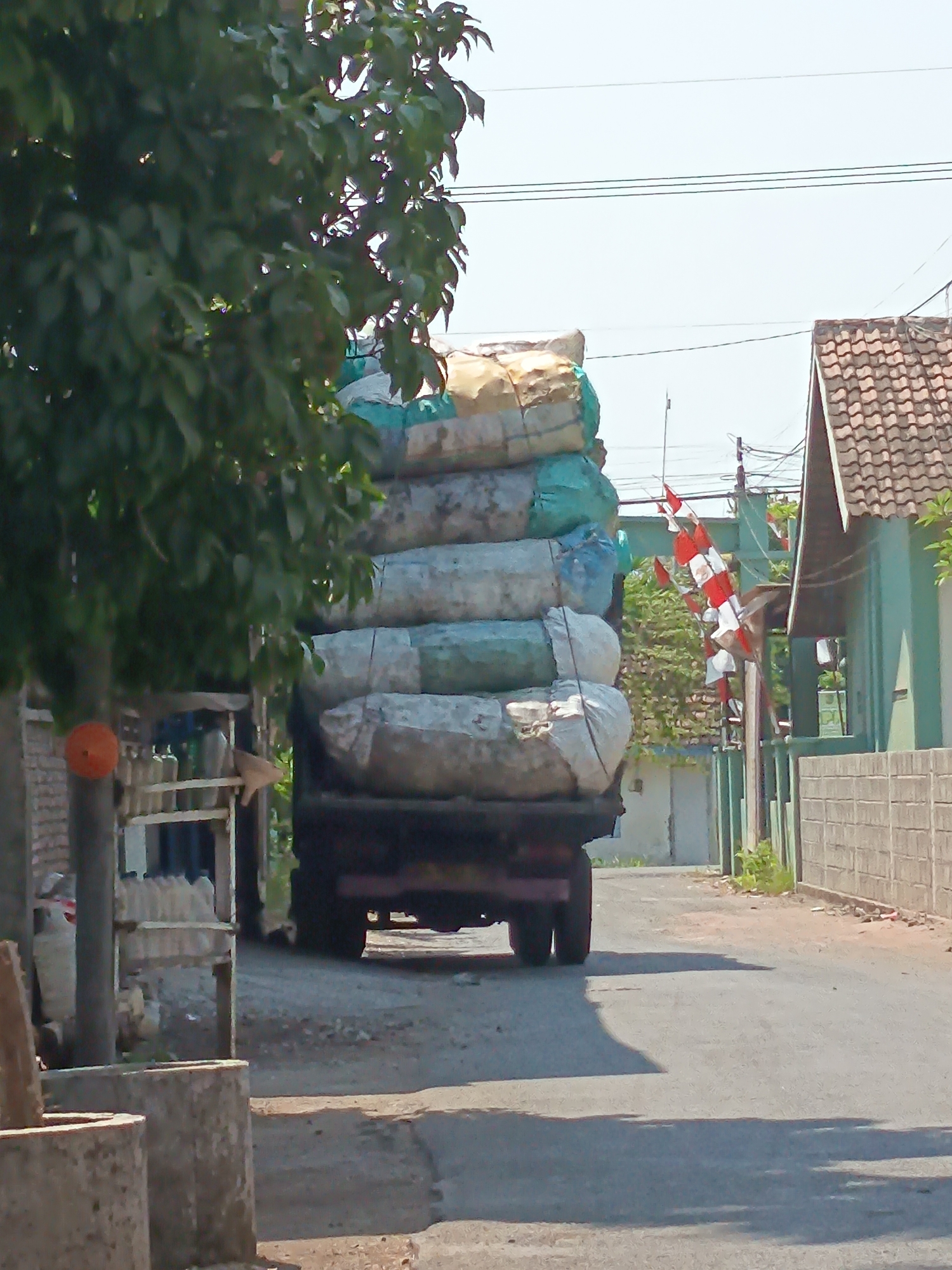 In the city of Kandangjati Wetan, Indonesia, a large truck is transporting a substantial load of bundled materials. The truck is seen from its rear, heading away from the camera down a paved road. The cargo is stacked high, composed of approximately six large cylindrical bundles wrapped in light-colored and teal fabric, secured with ropes. Some bundles appear to be made of straw or similar agricultural products.
To the left of the truck, a tree with dense green foliage partially obscures the view. Attached to the trunk of this tree is a bright orange spherical object, possibly a marker or decoration, and to the right of the tree trunk is a light-colored arrow-shaped object pointing towards the truck. Also on the left side of the road, a rudimentary structure with shelves holds several bottles or containers.
The environment appears to be a street in a residential or semi-rural area. On the right side of the road, a light green building with a tiled roof is visible. In front of this building is a section of a grey brick wall. Further down the street, several red and white Indonesian flags are strung vertically, possibly indicating a celebration or national holiday. The road surface is asphalt, with some gravel and debris scattered along the sides. The sky is clear and bright, suggesting a sunny day. The overall impression is a scene of everyday commerce or transport within a small Indonesian community.