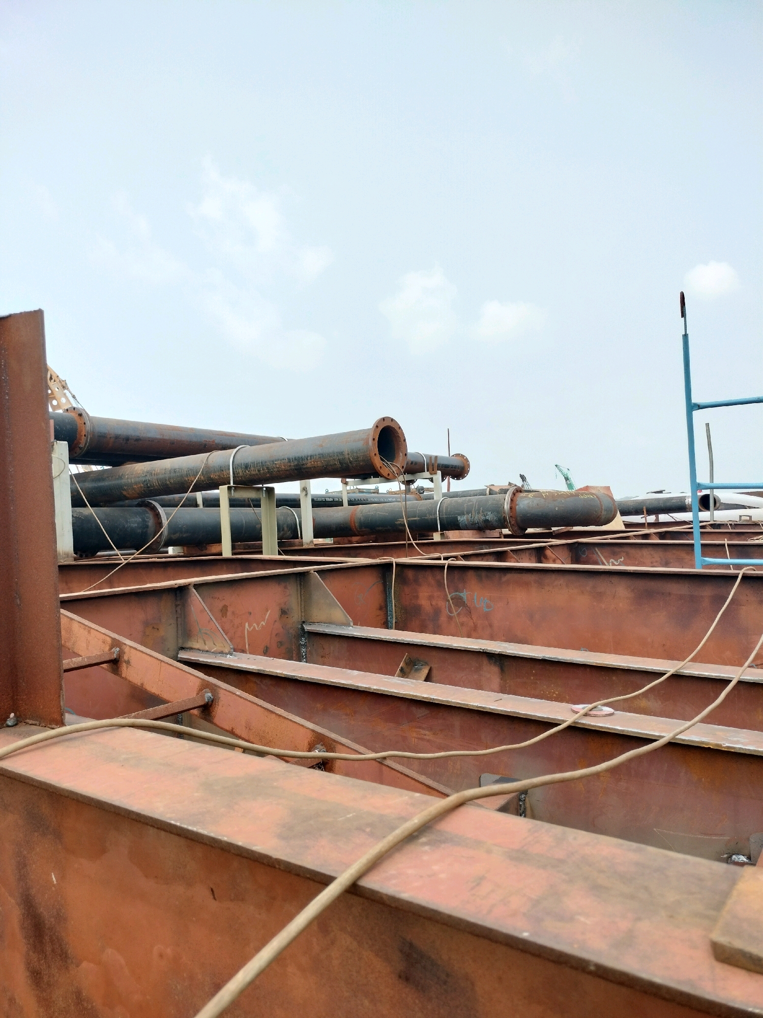 The image shows a construction site with large pipes and structural steel beams, likely in Batam, Indonesia, given the industrial setting. The sky is overcast with a few wispy clouds. Several large, dark, cylindrical pipes are arranged horizontally, supported by white bracing structures. These pipes appear to be made of metal and are of varying sizes.
In the foreground, there are large, reddish-brown steel beams, forming a complex structure. These beams have visible weld marks, indicating assembly work. A light-colored rope or cable is draped across these beams.
In the background, more pipes and steel structures are visible. A blue ladder-like structure can be seen on the right side of the image. In the far background, hints of industrial equipment and possibly cranes are present, suggesting ongoing heavy construction or shipbuilding activities. The overall scene depicts an industrial area undergoing construction or modification.