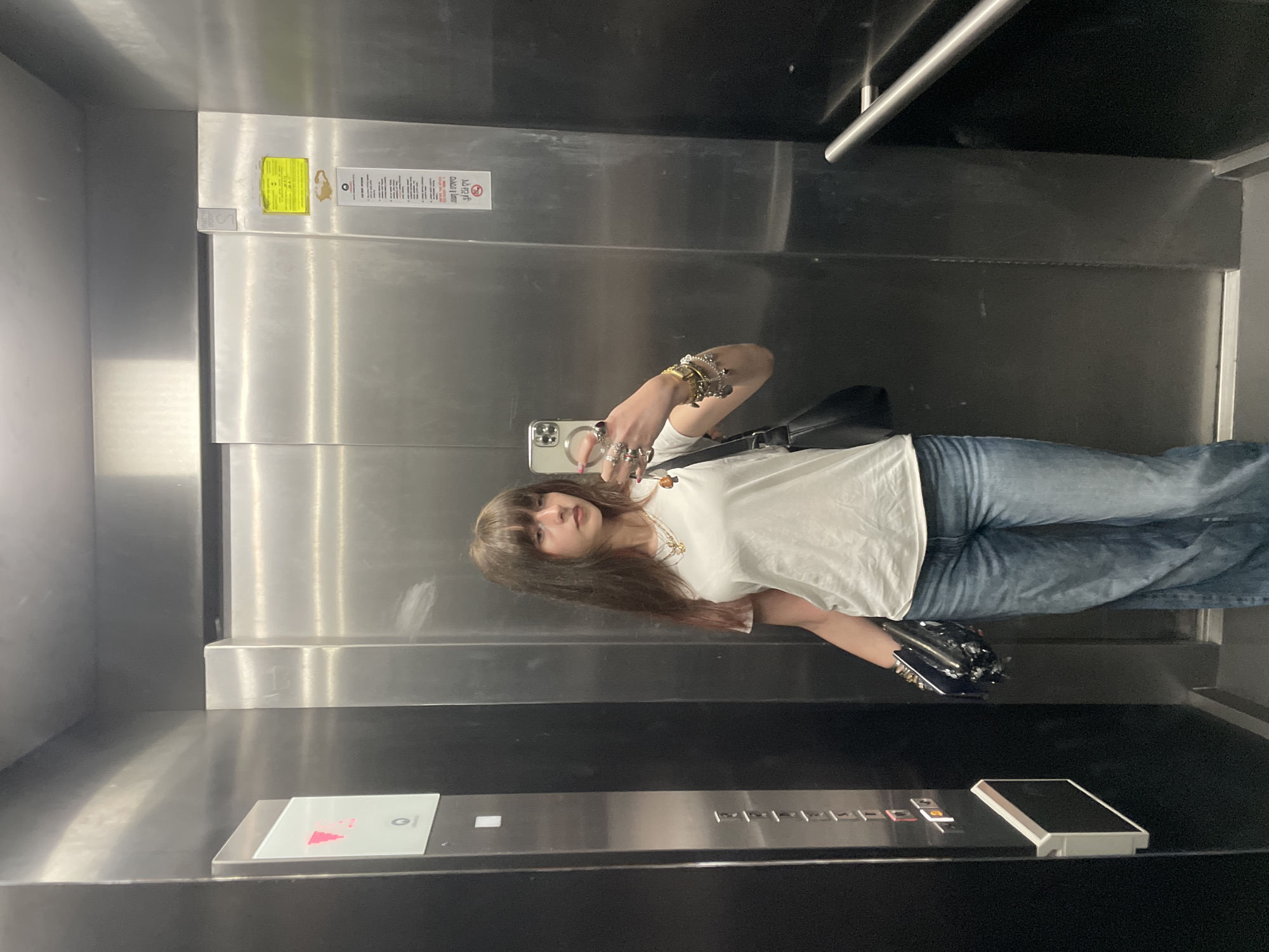 A person is taking a selfie in an elevator. The person has brown hair with bangs, is wearing a white t-shirt and blue jeans, and has multiple bracelets and rings. They are holding a phone in their right hand. Behind the person are stainless steel elevator doors. On the right side of the elevator is a control panel with buttons and a keypad. There are signs posted above the elevator doors. There is no evidence to indicate that this image was taken in Raanana, Israel.