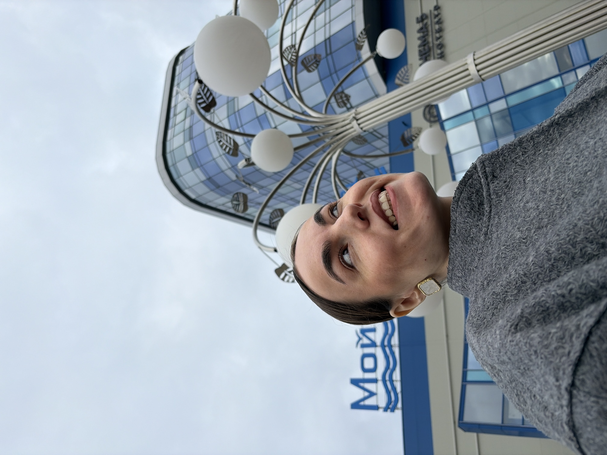 The image is a low-angle selfie of a woman with dark hair pulled back. She is smiling and looking up. She is wearing a gray sweater and square earrings. Behind her, the sky is overcast. In the background, there's an architectural detail consisting of a modern building with a blue and white grid pattern and a decorative lamp post with white spherical lights. In the lower left corner of the image, a sign in blue reads "Mой". This sign, combined with the architecture, indicates the location is likely the "Moy Theatre" in Izhevsk, Russia.