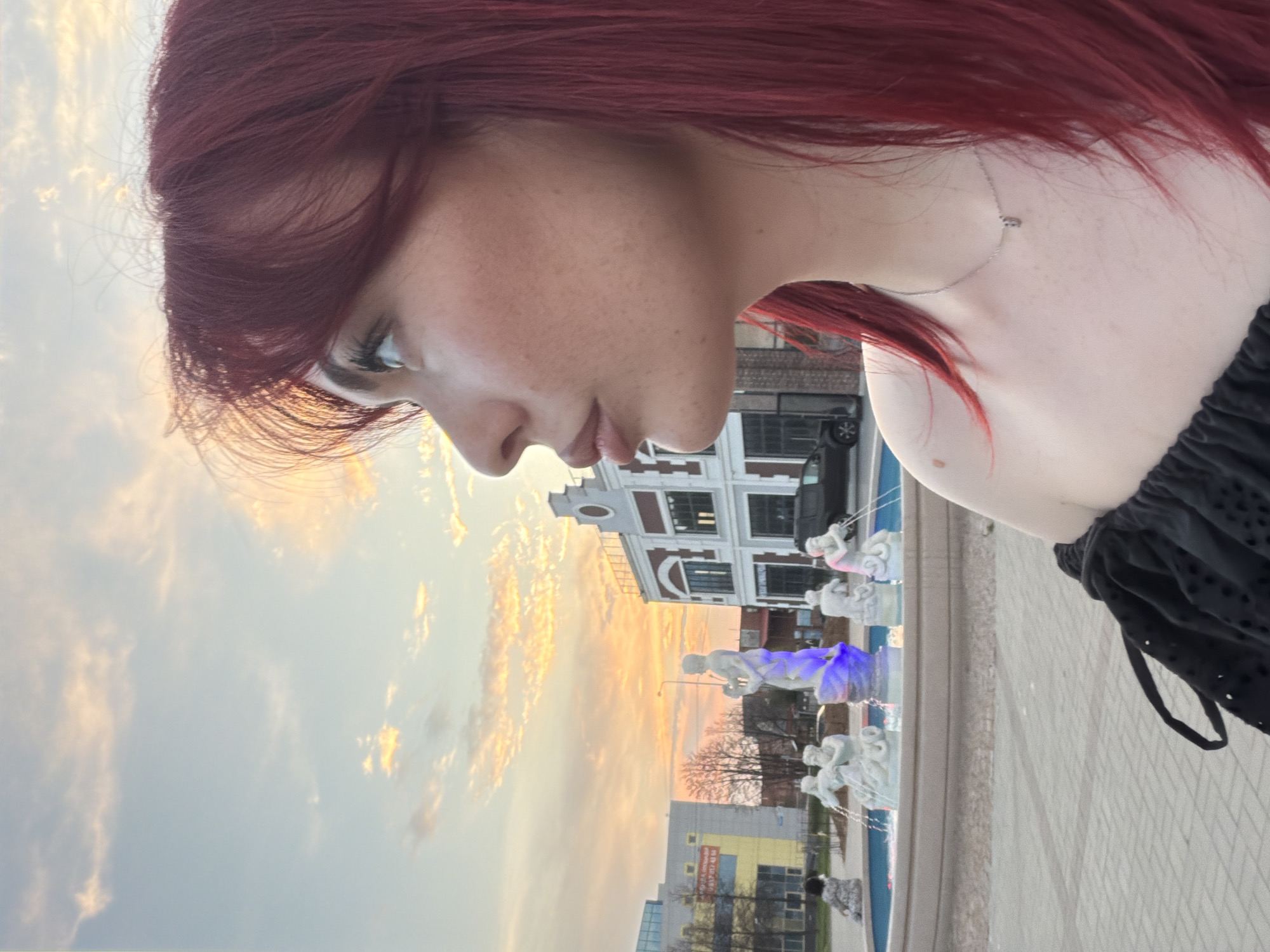 In Tolyatti, Russia, at sunset, a young woman with vibrant red hair is captured in a profile shot, looking towards the right side of the frame. She is wearing a black, off-the-shoulder top and a delicate silver necklace. In the background, a plaza features a multi-tiered fountain with white sculptures, some of which are illuminated with blue and pink lights. Beyond the fountain, buildings with modern and traditional architectural elements are visible, including a building with a yellow facade and another with red and white detailing. The sky is painted with the soft hues of a sunset, with scattered clouds catching the warm light. A black car is parked in front of one of the buildings.