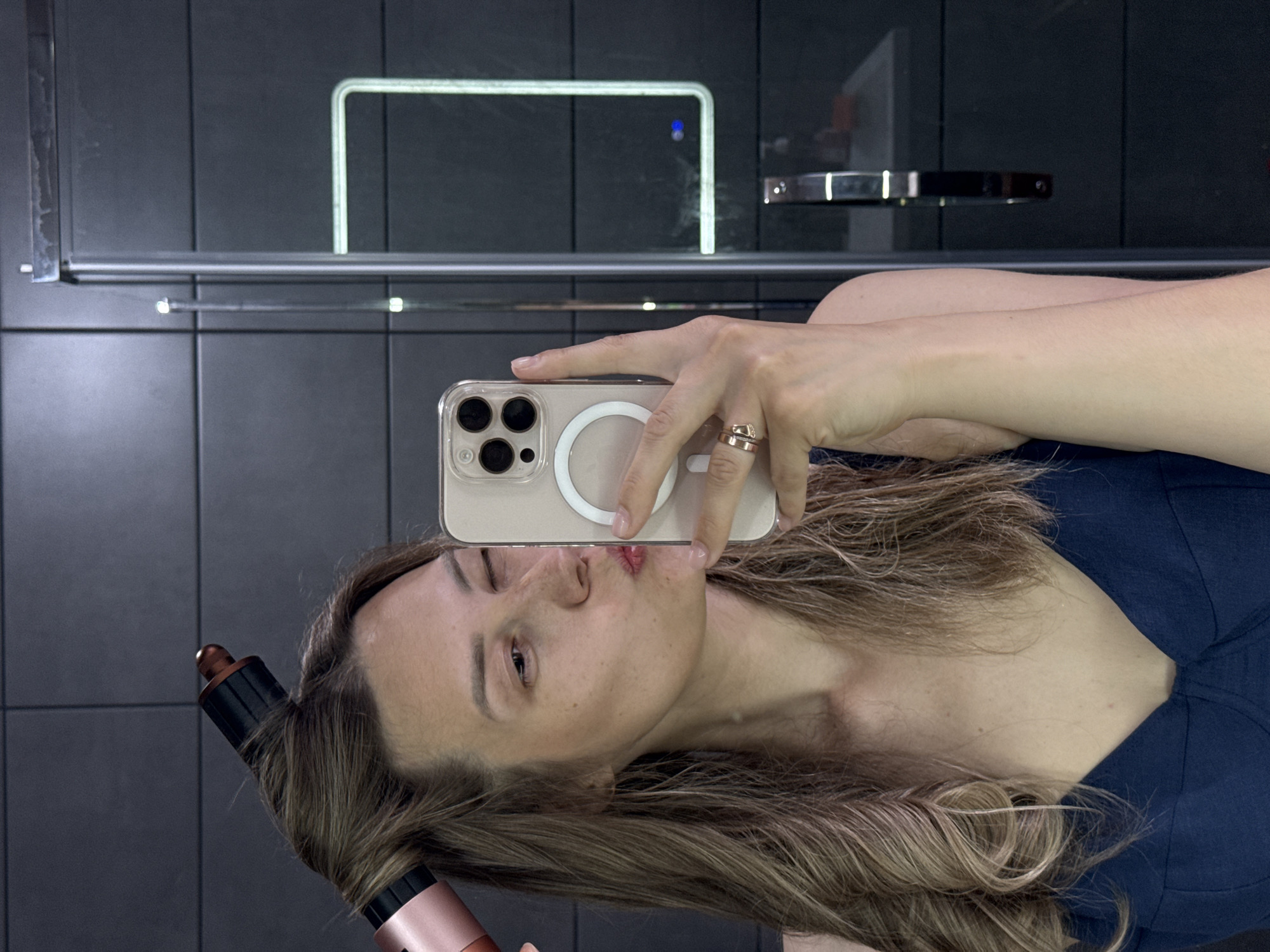 The image is a selfie taken by a woman with light brown hair in what appears to be a bathroom. She is holding a smartphone in her right hand, with a hair styling tool visible in her left hand near her hair. She is making a kissing face towards the camera. The background features dark gray tiles, a mirror with a built-in light, and a towel rack. The location in Chaikovsky, Russia is not discernible from the image.