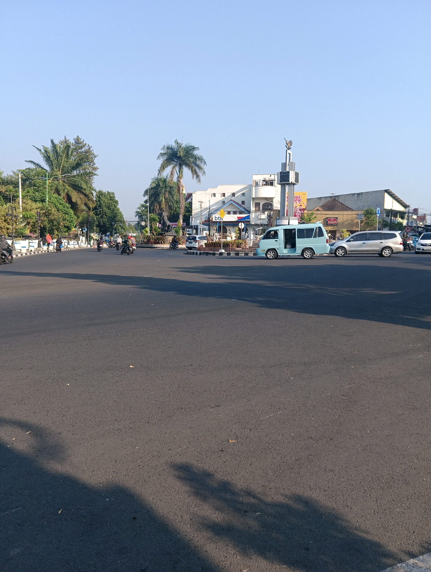 The image shows a street intersection in Salatiga, Indonesia, bathed in the soft light of what appears to be early morning or late afternoon. The sky is a clear, bright blue. In the foreground, a wide asphalt road dominates the frame, with shadows from unseen objects cast across its surface.
Several vehicles are present. A light blue minivan with its doors open is parked on the right side of the road. Beside it, a silver SUV is also stopped. Further down the road, a white sedan is visible. A person on a motorcycle is in the process of turning at the intersection, and other motorcycles are scattered throughout the scene, some moving and some parked.
The background features a variety of buildings, including a white, multi-story structure with a distinctive architecture and several other commercial buildings. A tall monument with a sculpture on top stands prominently in the distance, a landmark of the city. Trees, including palm trees, line the sides of the street, adding a touch of greenery to the urban landscape. The intersection appears to be a busy hub of activity, with a clear road marking indicating traffic flow.