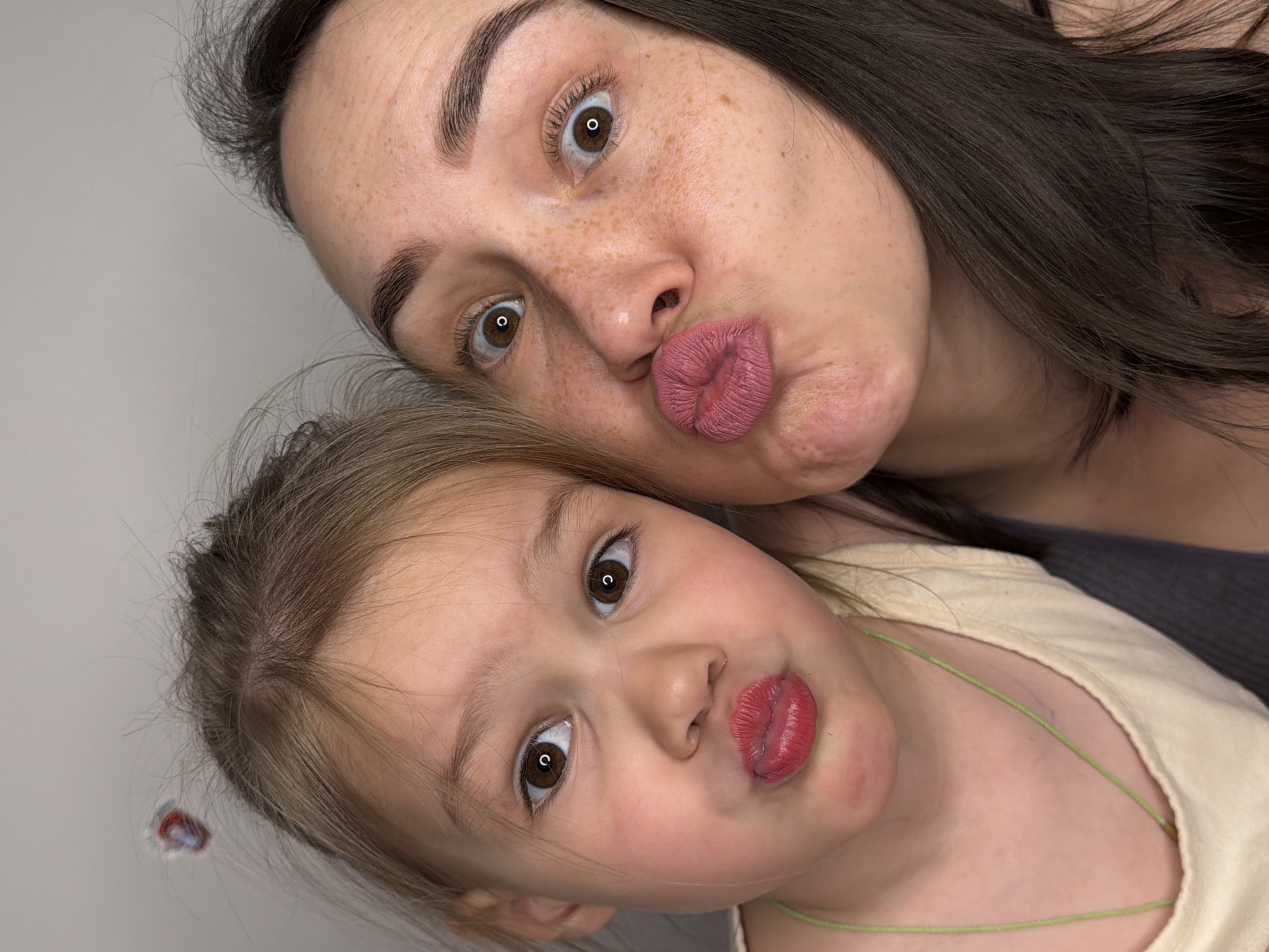 The image shows a close-up of a woman and a young child. Both are making a "kissing" face at the camera. The woman has dark hair and freckles. The child has light brown hair. The background is a plain, light gray or white. There is no visual evidence to confirm the location is Chaikovsky, Russia.