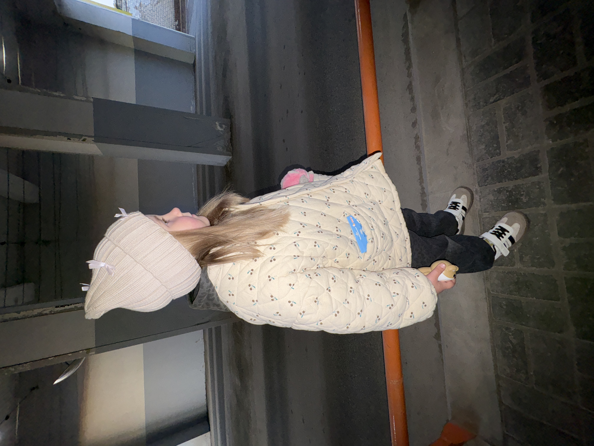 A young girl in a light-colored jacket and a knit hat is standing in what appears to be an alleyway in Chaikovsky, Russia. She is holding a small stuffed animal. The alleyway has brick paving and is bordered by concrete structures. An orange pipe runs vertically behind the girl. The overall lighting in the alleyway is dim.