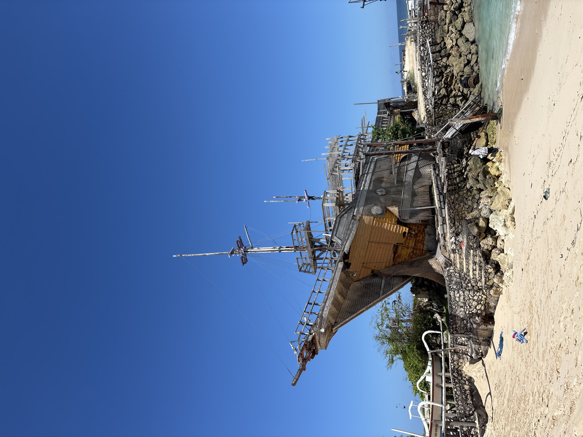 The image depicts a weathered, partly wooden ship structure resting on a rocky outcrop adjacent to a beach. The sky is clear blue, and the beach has light-colored sand. The ship's frame is visible, with remnants of masts and rigging. The location is near the ocean, where gentle waves meet the shore. Evidence of built structures, including what appear to be pathways and possibly other architectural elements, are visible surrounding the ship. The scene's location can be identified as Pantai Melasti in Kuta Selatan, Indonesia. A few items that appear to be clothing, towels, or plastic containers are scattered on the sand. The overall environment suggests a tourist attraction or a site with some historical or recreational significance.
