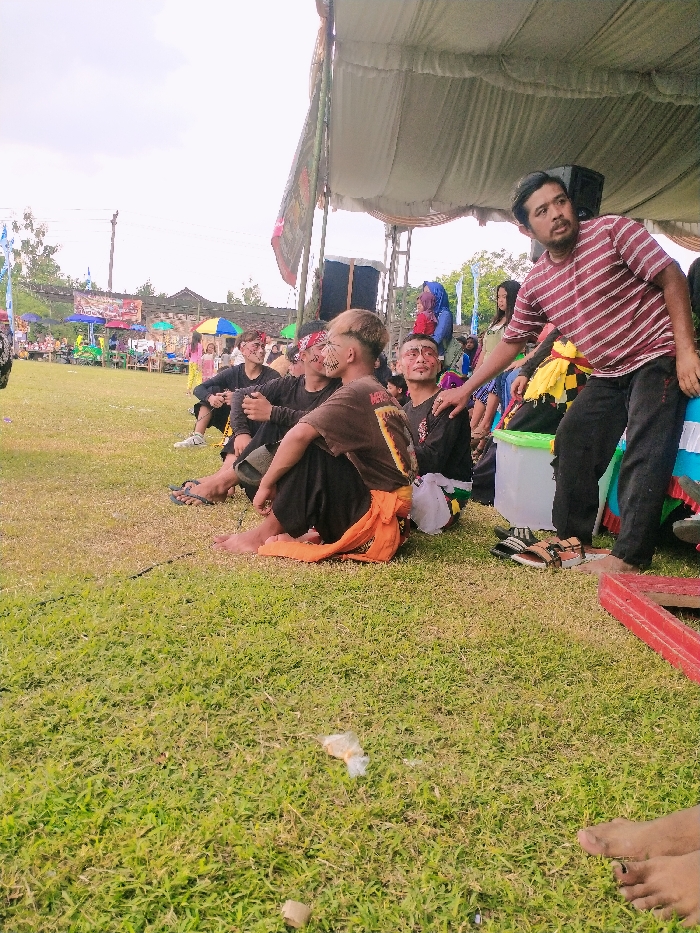 The scene is a grassy field in Karangmalang, Indonesia (latitude: -7.47, longitude: 111.064). There are several men sitting on the grass under a large white canopy tent. They are wearing casual clothing, some with colorful face paint. There are two men sitting on the grass behind the group, also wearing casual clothing. Some are wearing traditional Indonesian clothing. Some men are holding food and drinks, and others are chatting. The environment is bright and sunny, with green grass and a few trees in the background. The men are smiling and laughing. There is a red box on the grass near the group. A white plastic container sits next to the group, with the side missing. There is also a large speaker located behind the tent, but it is not visible in the image.