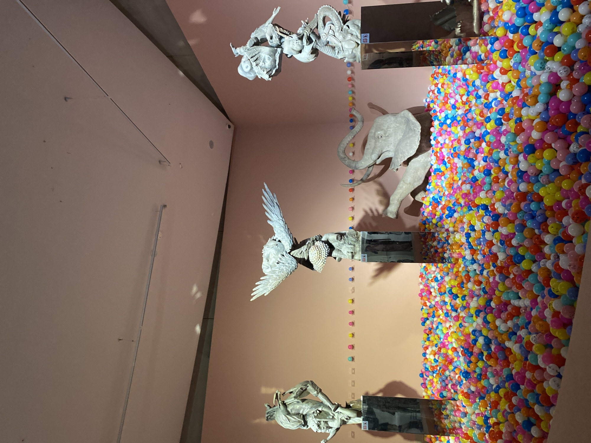 This image depicts an art installation, likely in a museum or gallery, featuring a vibrant ball pit filled with numerous colorful plastic balls. In the background, several sculptures are displayed on pedestals against a peach-colored wall. To the left, a sculpture of a person or mythical creature stands on a dark, reflective pedestal. In the center, a sculpture of a winged creature, possibly an eagle or phoenix adorned with pearls, is positioned above a smaller sculpture. Further to the right, an elephant sculpture is partially submerged in the ball pit, with its trunk and head visible. The right side of the scene features another intricate sculpture, a multi-layered figure with various mythical elements, also on a dark pedestal. A line of small, colorful spheres or objects is arranged horizontally on the wall behind the elephant sculpture. The lighting appears to be from above, casting shadows. The context of Jinniu District, China is not visually evident from the image itself.