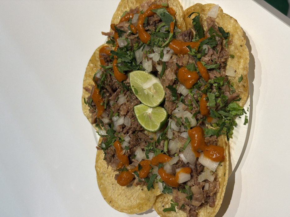 The image displays five tacos arranged circularly on a white plate. Each taco features a golden-brown corn tortilla holding shredded dark meat. The meat is topped with finely diced white onion, chopped green cilantro, and an orange-red sauce applied in distinct drizzles. Two bright green lime halves are positioned centrally atop the assembly. The plate rests on a smooth, white surface, likely a table or counter, against an undecorated white background, suggesting an indoor environment. No individuals, actions, or specific interactions are depicted. The image contains no visual elements that identify its location as Shibuya, Japan, or any particular city.