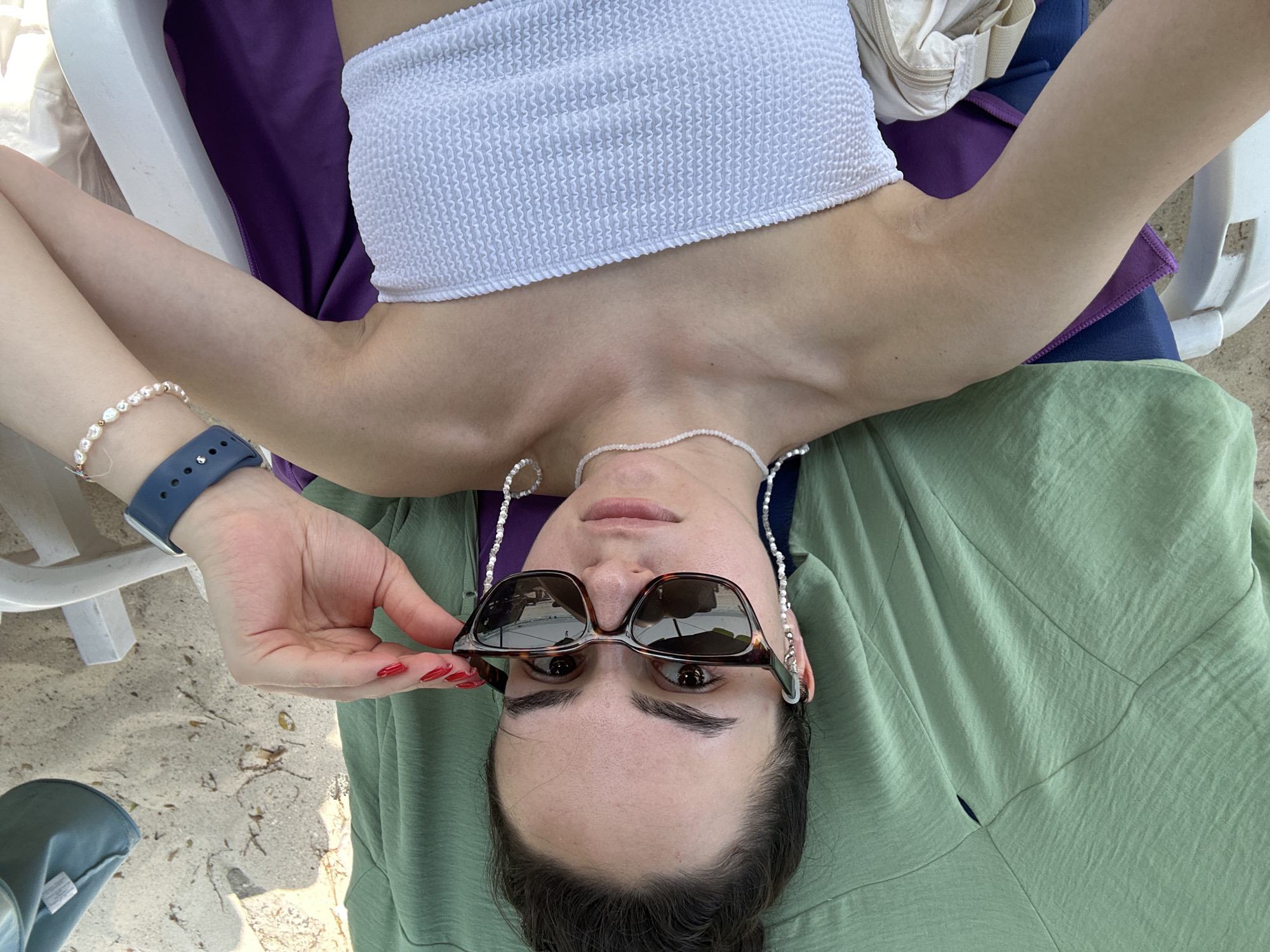 A woman lies supine outdoors, likely on a lounger. She wears a white, textured bandeau top and an olive green garment. Her arms are raised, one holding tortoiseshell sunglasses with a beaded chain. She wears a blue smartwatch and a pearl bracelet on her left wrist, and a delicate pearl necklace around her neck. A purple towel is visible behind her upper body. The background suggests a sandy area, potentially a beach. The photograph appears to be taken from a high angle, looking down at the woman. The location is possibly Pattaya City, Thailand.