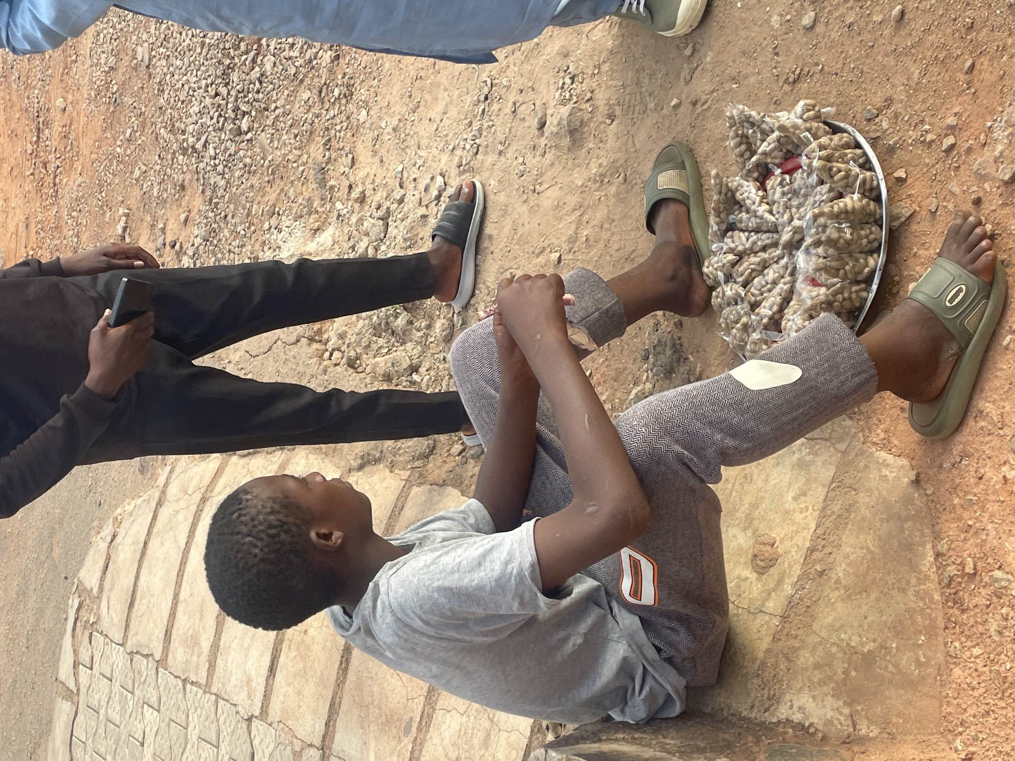 The image shows three people in an outdoor setting in Jos, Nigeria. A young boy is seated on what appears to be a stone or concrete structure, holding a metal tray filled with small, packaged goods - likely peanuts or snacks. He is wearing a grey t-shirt, patterned grey pants, and green sandals. To the boy's left, another individual is standing, holding a mobile phone. They are wearing black pants and a dark shirt. Partially visible in the upper part of the image is a third person wearing jeans and sneakers. The ground is a mix of dirt, gravel, and paved areas, suggesting a semi-urban or transitional environment. The image captures a casual, everyday scene, possibly depicting the boy selling snacks.