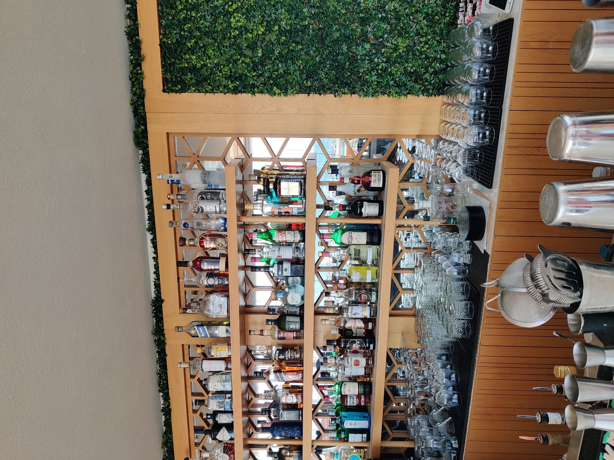The image shows a well-stocked bar with shelves full of liquor bottles. The shelves are made of wood and are arranged in a grid pattern. A large section of the wall to the right is covered with artificial greenery. In front of the shelves, there are many glasses, neatly arranged and ready for use. The bar counter below has various bar tools, including shakers and strainers. The lighting is bright, suggesting it is daytime. The overall scene depicts a clean and organized bar, possibly in a restaurant or club.