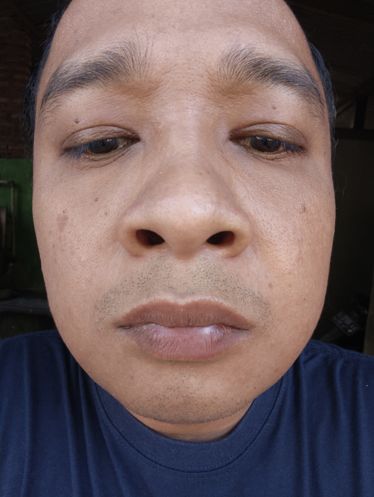 A close-up, front-facing photograph shows the face of an adult male. The individual is looking directly at the camera with a neutral expression. The image focuses on the upper portion of his face, from just above the eyebrows down to the collarbone. The lighting suggests an outdoor setting, possibly during the day, with natural light illuminating the subject's face. The background is blurred, showing hints of greenery and possibly a building structure, but it is not detailed enough to identify a specific location within Kaliwader, Indonesia. The man has dark hair and eyebrows, and his skin tone is medium to dark. He is wearing a dark blue t-shirt, the neckline of which is visible at the bottom of the frame. The photograph is taken from a slightly low angle, emphasizing the subject's features.