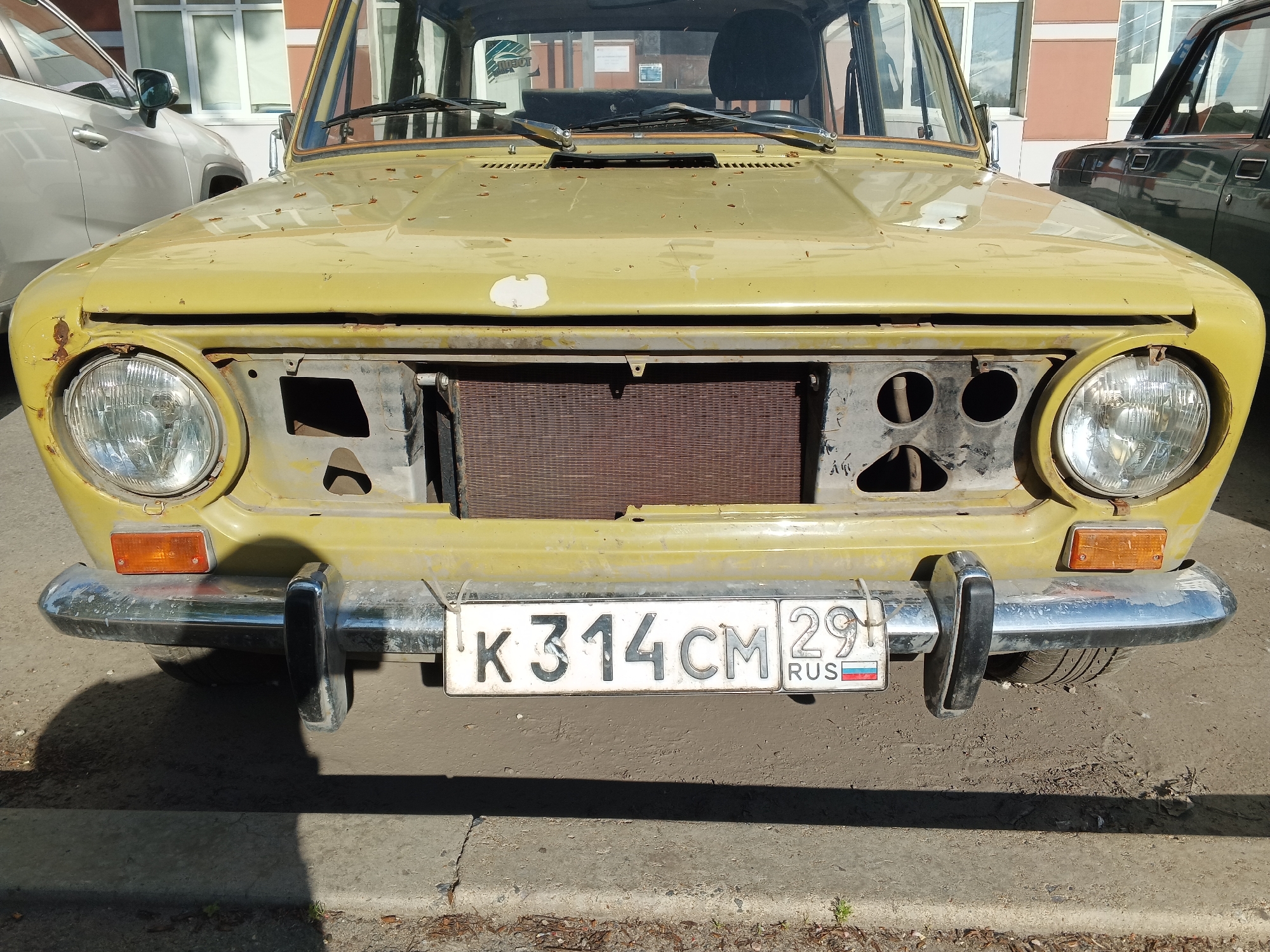 The image shows the front of a yellow, older model car parked on a paved surface. The car has a missing grille, revealing the radiator. The license plate, K 314 CM 29, is visible and reads "RUS" with the Russian flag. The car appears to be in a state of disrepair, with visible rust and peeling paint. In the background, a light-colored SUV is parked to the left, and a darker vehicle is parked to the right. A building with windows is also visible behind the cars. The scene appears to be in an urban environment, consistent with a location in Arkhangelsk, Russia, given the license plate information. The lighting suggests it is daytime. No people are visible in the image.