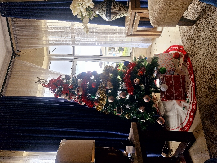 A festive Christmas tree, adorned with colorful ornaments and tinsel, stands near a window. Presents wrapped in festive paper sit beneath the tree on a white and red tree skirt. Blue curtains frame the window and tree. The scene is set on a tiled floor. A table with a vase of white flowers and a woven decoration sits near the window and a chair is visible in the background.