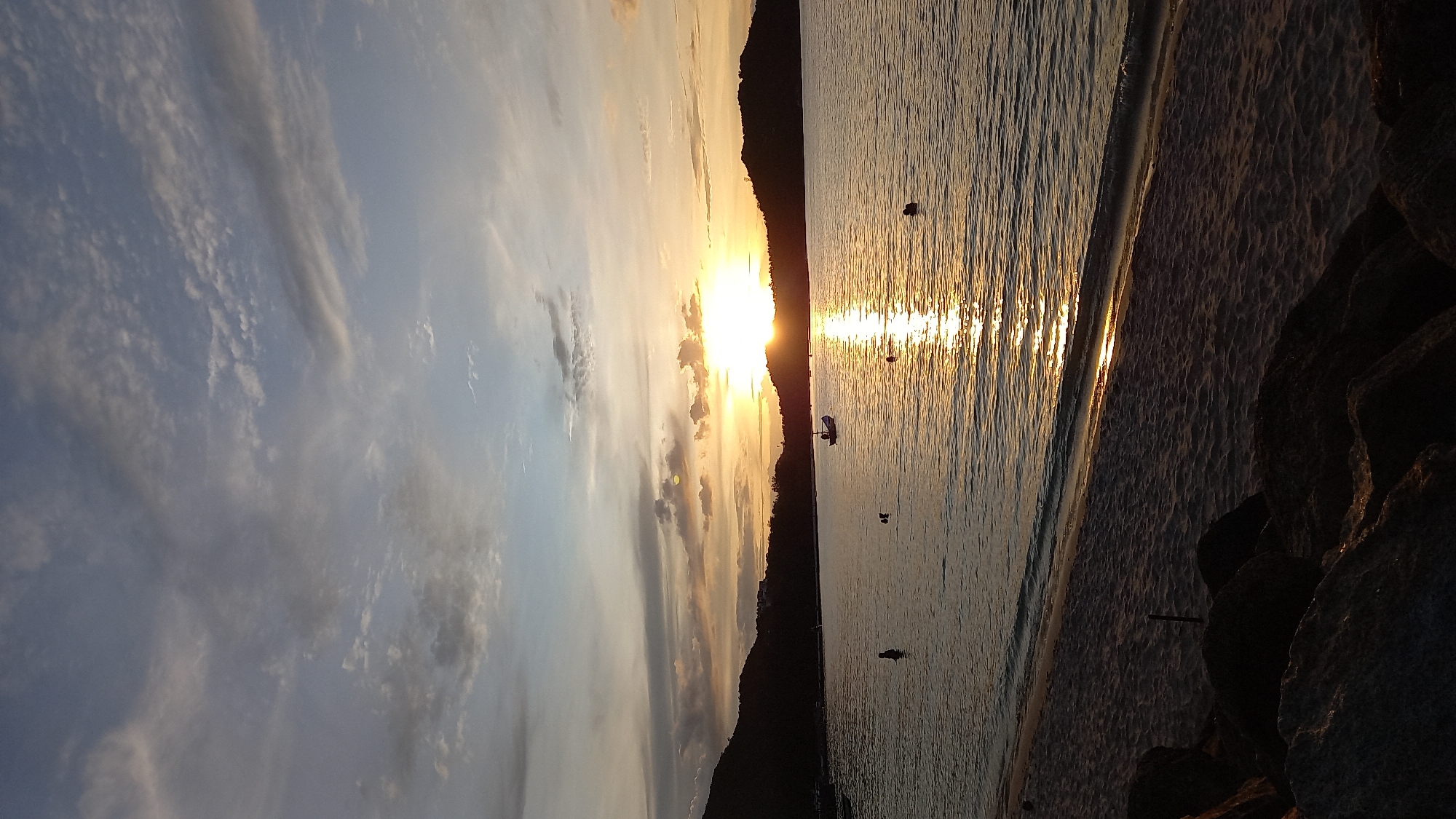 This image captures a serene sunset scene at Koh Phangan, Thailand. The sky is filled with a mix of wispy clouds, transitioning from a pale blue to hues of orange and yellow as the sun dips below the horizon, silhouetting a dark mountain range. The sun's reflection casts a shimmering golden path across the calm water, creating a peaceful and tranquil atmosphere.
Several small objects, possibly buoys or markers, are scattered in the water. A lone figure can be seen wading in the shallow water near the beach, while another small boat with a canopy is anchored further out. The beach itself is made of sand, with a line of dark, rough rocks forming the foreground on the right side of the image. The overall mood is one of quiet beauty and relaxation, typical of a tropical evening. The time of day is clearly sunset, indicated by the sun's low position and the warm, golden light. The weather appears calm and clear, with only a few scattered clouds. There is no visible text in the image.