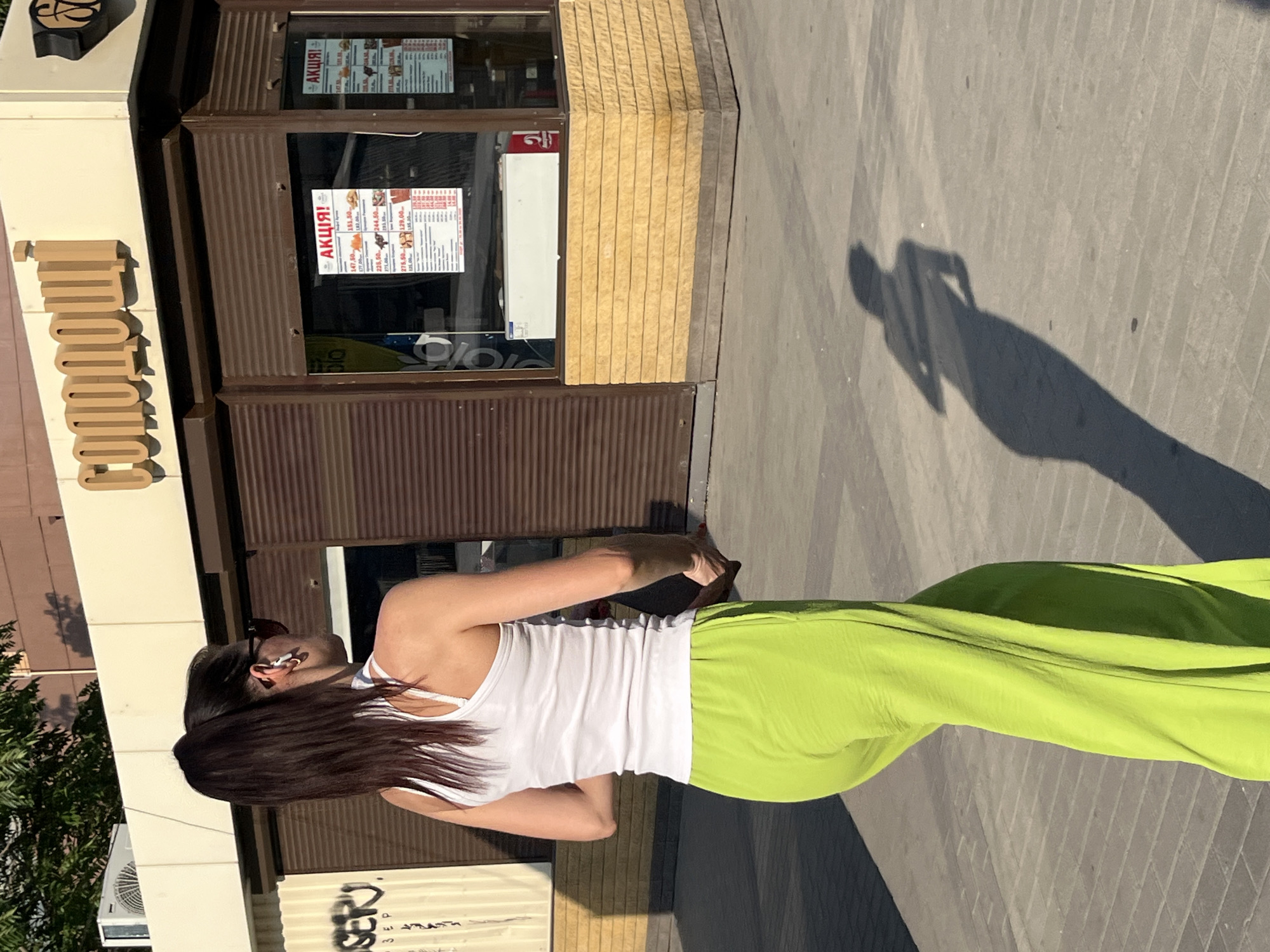 The image shows a woman from behind, wearing a white tank top and bright green pants, walking on a sidewalk. She has long, dark hair and is wearing sunglasses and earrings. A shadow of the woman is cast on the pavement, indicating it is daytime, likely sunny. The background features a building with a storefront that has signs indicating "АКЦІЯ!" (Action! or Sale! in Ukrainian). The building has a sign with large, brown, block letters spelling "CLOUDY". There is also an air conditioning unit visible on the side of the building, and some graffiti on a lower section. The overall setting appears to be a commercial street or area in Dnipro, Ukraine. The woman is walking away from the camera, and her posture suggests a casual stroll.