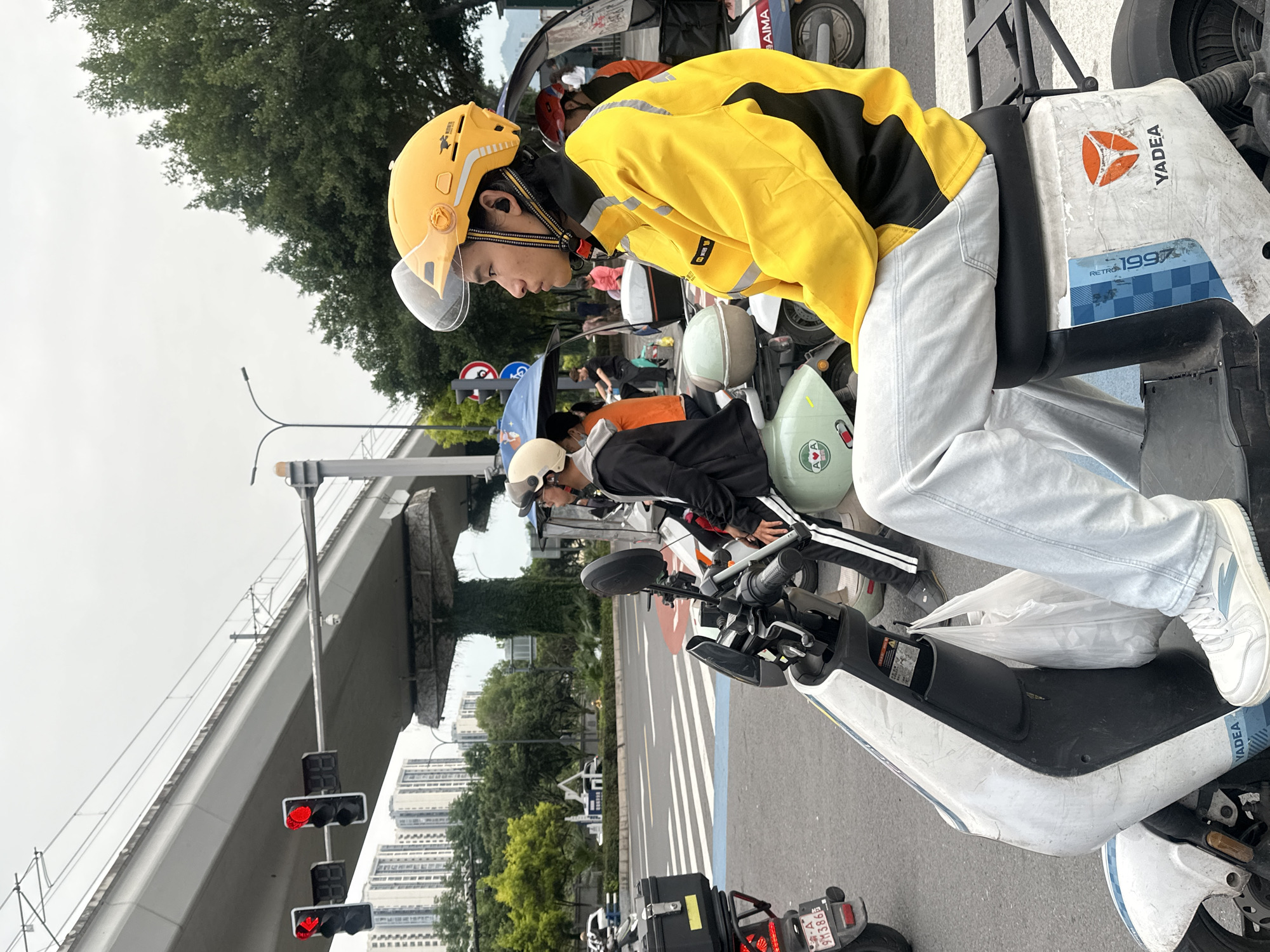 This is a street-level photograph taken in the Chongqing High-tech Zone, China, depicting several individuals on electric scooters waiting at a traffic light. In the foreground, a young man wearing a yellow jacket and helmet is seated on a white and blue electric scooter, facing to the right. His left hand is on the scooter's handlebars, and his right hand rests on his leg. He is wearing light-colored pants and white sneakers. In the background, another person in a black jacket and white helmet is visible on a scooter. Behind them, several other scooters and people are present, some under canopies. Traffic lights displaying red signals are visible overhead, attached to a large, elevated concrete structure, likely an overpass, with power lines running above it. The environment includes trees and tall buildings in the distance. The overall scene is one of urban transit and waiting.