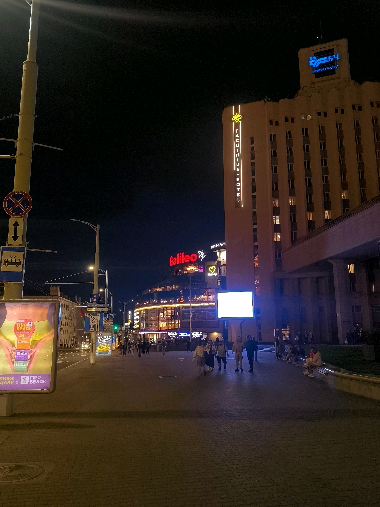 The image depicts an urban street scene at night in Minsk, Belarus. Dominating the right side of the frame is a tall, light-colored building, possibly a hotel, with illuminated windows. A prominent sign on the building reads "ГЯСНIЧНА • HOTEI". At the very top of this building, a large blue digital display shows "БЧ" and "www.rw.by".
On the left side of the street, a large billboard with vibrant graphics displays advertisements, including "ПРО БЕЛОК". There are also street signs, one featuring a "no parking" symbol and another indicating a taxi. The street is paved with cobblestones and illuminated by streetlights and the glow from building signs and the digital display.
Several people are visible walking along the sidewalk and on the street, appearing to be engaged in typical evening urban activities. Some individuals are seated on a low wall or bench on the right side of the image. The overall atmosphere suggests a bustling city center after dark, with commercial establishments and public spaces illuminated. The presence of the "www.rw.by" website suggests a connection to Belarusian Railways.