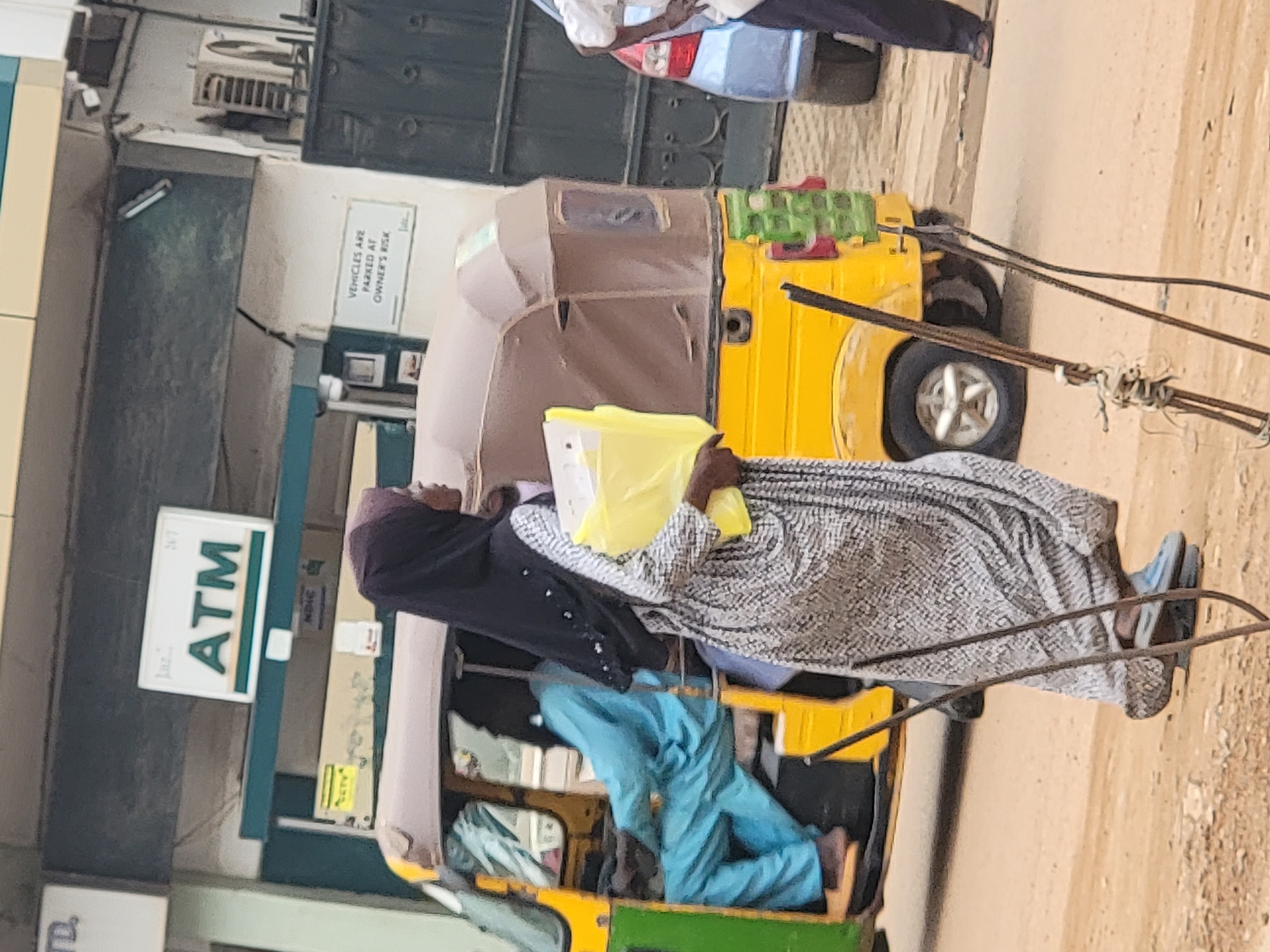A man in Maiburiji, Nigeria, stands near a yellow auto rickshaw in front of an ATM. The man wears a head covering, a grey patterned shirt, and blue pants. He holds a yellow object. Another person is in the rickshaw. Vehicles are parked in the background.