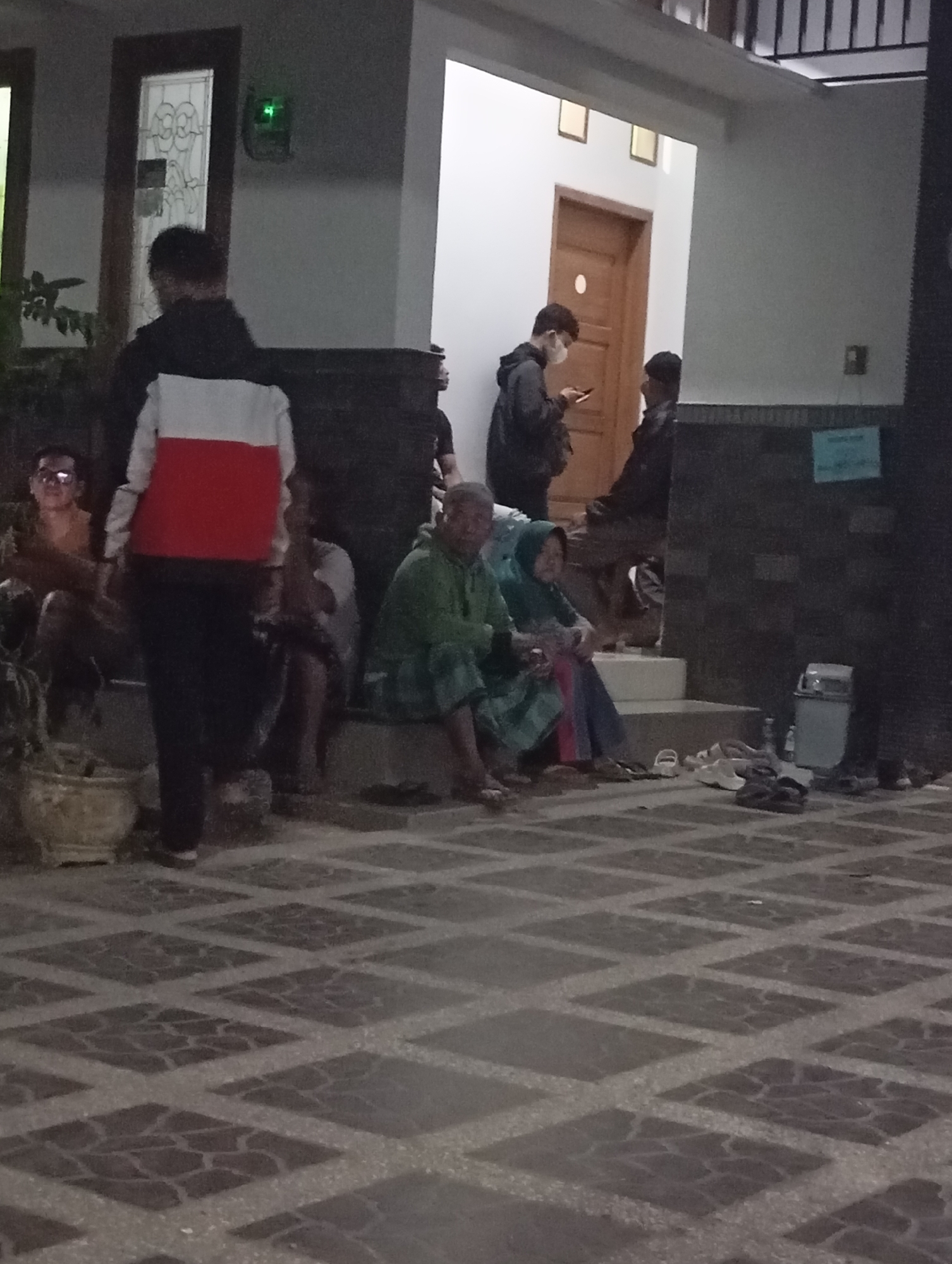 The image depicts a nighttime scene on the tiled patio of a residential building in Banjarsari, Indonesia. Several individuals are present. In the foreground, a person stands wearing a jacket with red and white horizontal stripes. To the left, a man with glasses is seated, appearing to be in conversation with another individual partially visible. Further to the right, three people are seated on steps. One, wearing a green garment, is looking away from the camera. Another, a woman in a hijab, is seated next to him. The third seated individual is largely obscured. In the background, two young men are standing near a doorway. One, wearing a face mask, is looking at his phone, while the other stands beside him. A large plant pot is situated on the left side of the patio. A trash receptacle and several pairs of sandals are visible on the ground. The tiled floor features a diamond pattern.
