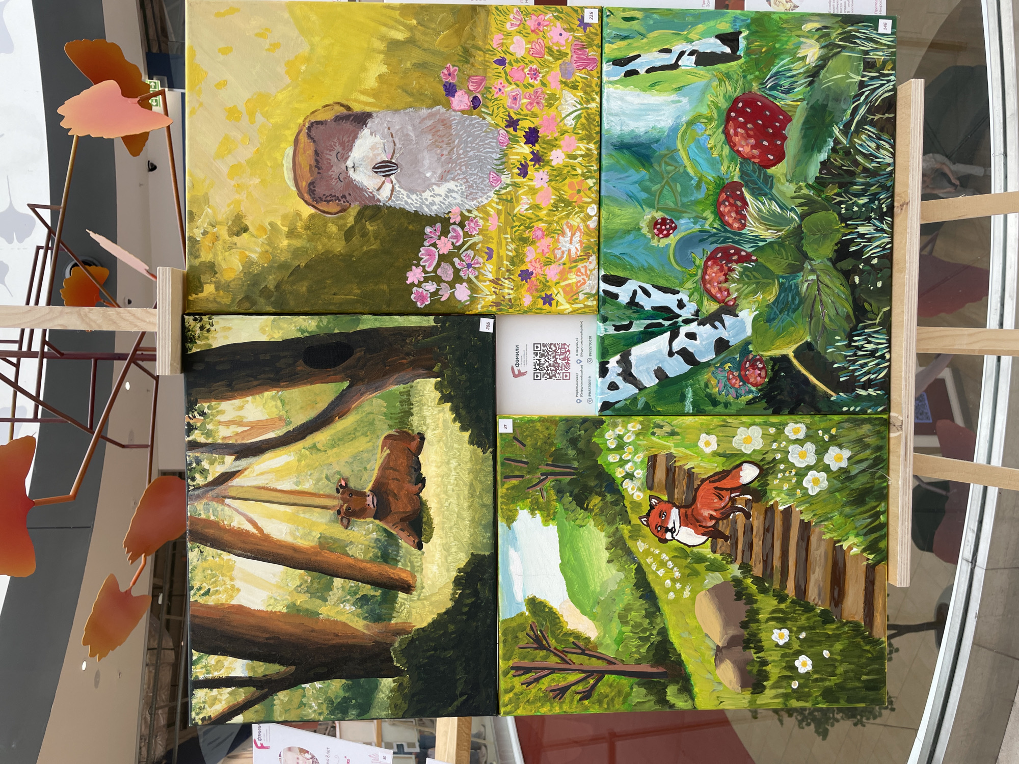 This media file features a collection of four acrylic paintings displayed on wooden easels. The paintings appear to be part of an exhibition or art sale, given the presence of QR codes and text that seems to be related to the artist or venue.
The subjects of the paintings are varied:
1. The top left painting depicts a fluffy, grey and brown creature, possibly a hamster or guinea pig, with a neutral expression, nestled amongst yellow and green foliage with small pink and purple flowers.
2. The top right painting showcases a vibrant scene of bright red strawberries growing on green vines, interspersed with abstract black and white shapes that could represent water or a stylized background.
3. The bottom left painting presents a serene forest scene with dappled sunlight filtering through trees. A brown cow with a gentle expression is lying down amidst the greenery.
4. The bottom right painting features a cheerful red fox with a white chest and tail, lying on a path made of wooden planks. The scene is lush with green foliage and small white flowers.
The overall setting seems to be an indoor space, possibly a gallery or a public area, with large windows or glass doors allowing natural light to enter, as indicated by reflections and the ambient lighting. The metal decorative elements on the left suggest an artistic or modern interior design.
Visible text includes what appears to be Russian, with phrases like "Фэмили" (Family), a street address "В.Засулич 42", and phone numbers. A QR code is also visible on a card attached to one of the paintings, likely for more information. The presence of numbers like "226", "286", "146", and "87" near the paintings might indicate item numbers or artist identifiers. The phrase "8 лет" (8 years) is also partially visible, possibly referring to the age of the artist or the duration of an event.