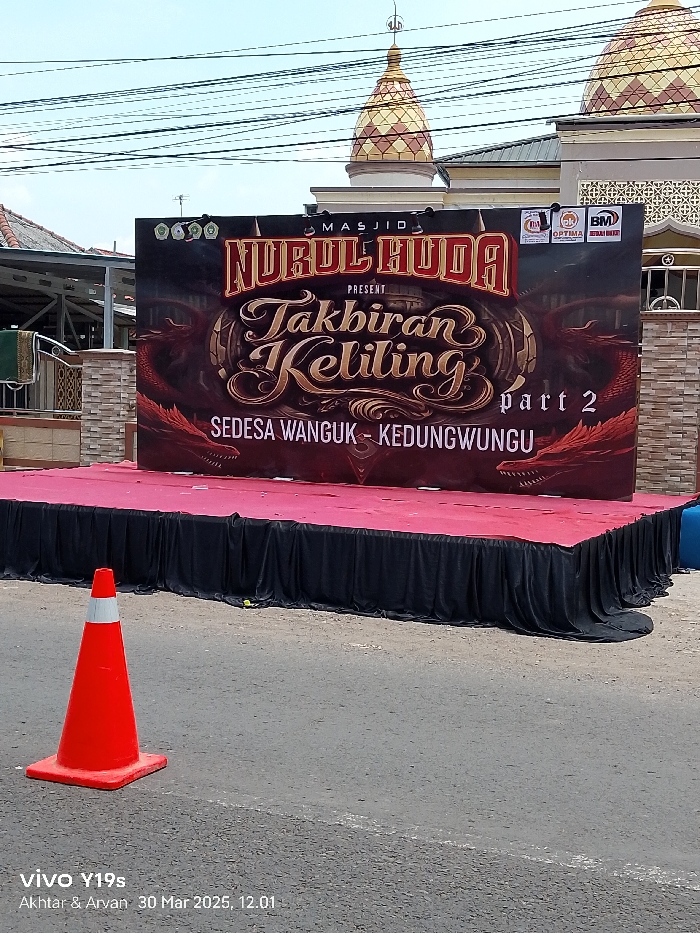 A black stage draped with a red fabric cover is set up in front of a large sign in Anjatan, Indonesia (latitude: -6.41, longitude: 107.961). The sign advertises an event called "Takbiran Keliling Part 2" hosted by Masjid Nurul Huda. The sign features a dragon with a red, orange, and yellow color scheme. A single orange traffic cone stands in the foreground. The background features a partially visible mosque building. The area appears to be a street. The sign is surrounded by electrical wires strung overhead. A second sign is partially visible to the right, and the remnants of a blue object are visible in the bottom right corner.
