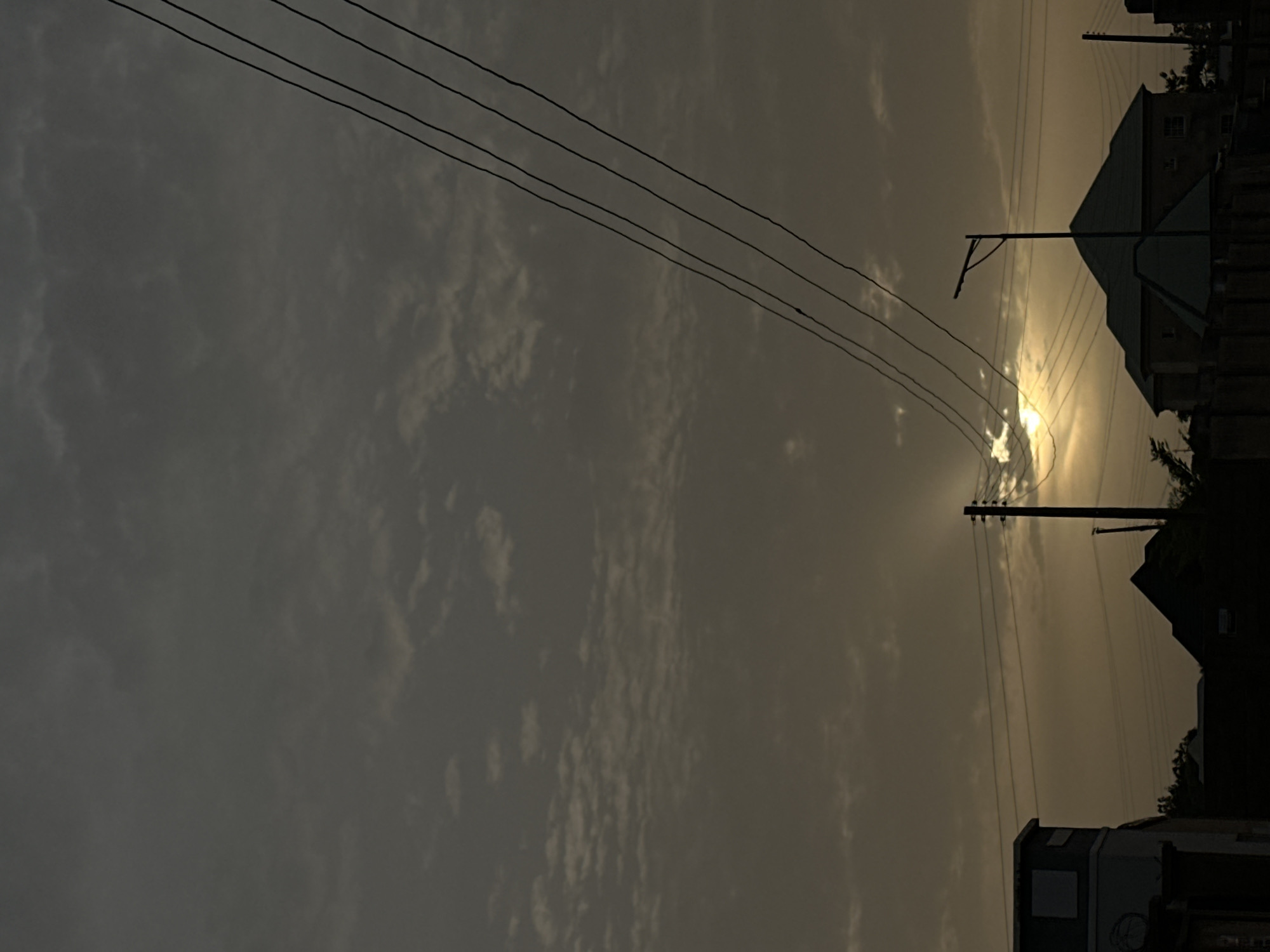 The image shows a landscape at dusk or dawn, with buildings and electrical infrastructure in Kirana, Nigeria. The sky is overcast, with the sun obscured by clouds, creating a diffused, muted light. Several power lines stretch across the upper portion of the frame. Utility poles are visible amidst the buildings. In the foreground, a vehicle can be seen.