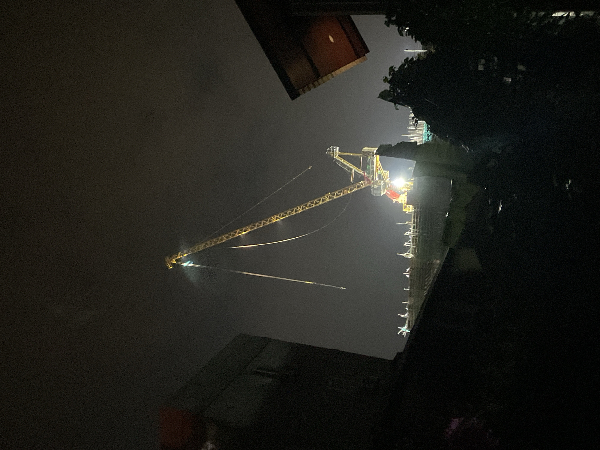 An elevated construction crane with bright yellow and red components is illuminated by lights against a dark, cloudy night sky. The crane's boom extends diagonally upward and to the left, with long, thin cables hanging from it. Below the crane, a section of a building or structure under construction is visible, also lit by artificial lights. In the foreground and on the sides of the image, the dark silhouettes of buildings, trees, and foliage obscure the lower portions of the scene. The image appears to be taken from a residential area overlooking a construction site. The location is identified as Phu Thuy Ward, Vietnam.