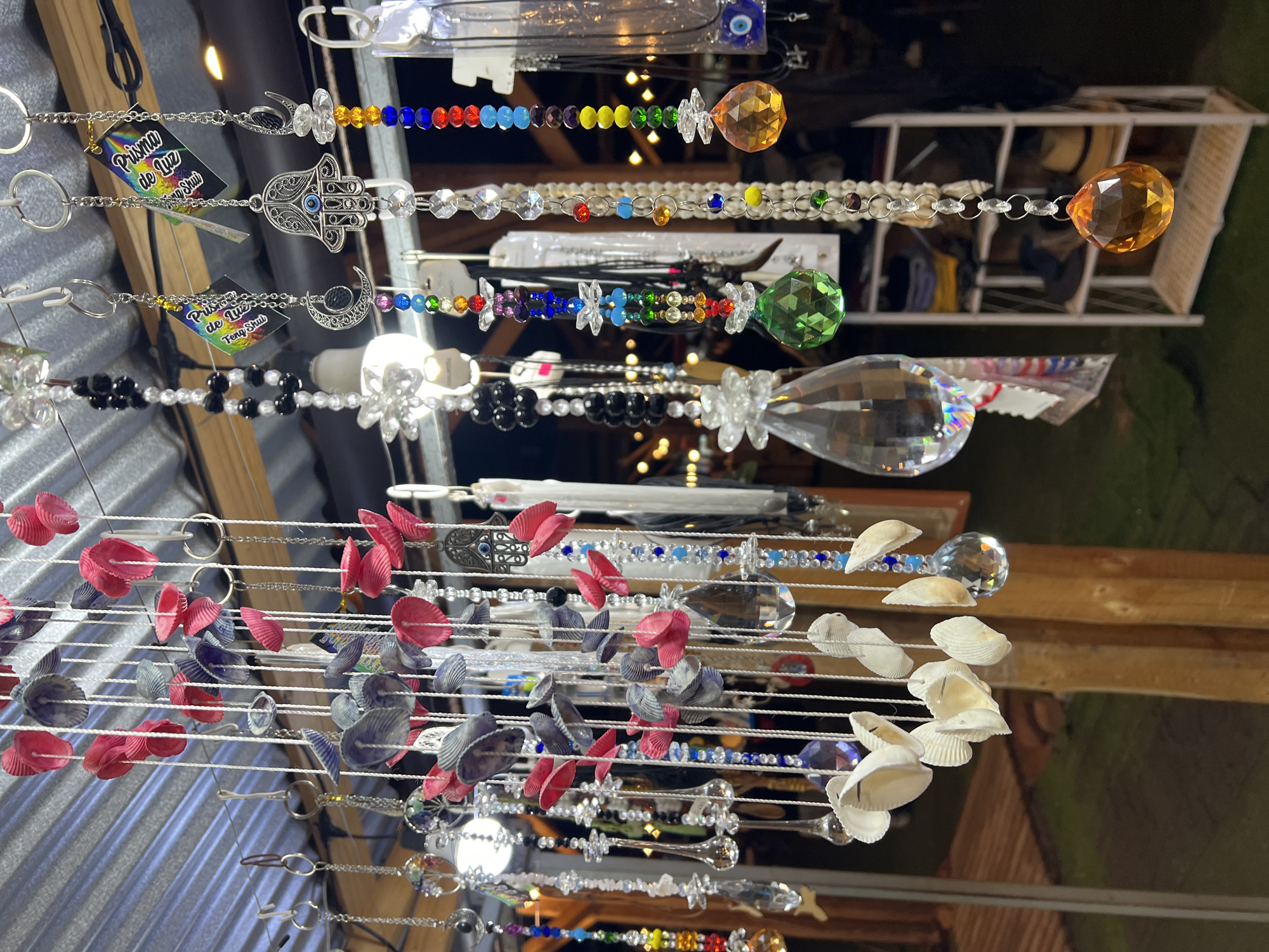 This image depicts a collection of decorative wind chimes and hanging ornaments displayed outdoors, likely at a market or street stall in Arica, Chile. The items are suspended from a corrugated metal roof.
The ornaments are diverse, featuring various materials and designs. Some are made of small, colorful seashells in pink, purple, and white. Others incorporate glass beads in a rainbow spectrum, clear crystal pendants of different shapes and sizes, and charms such as crescents, Hamsa hands with an evil eye, and circular motifs. Several items have small tags attached.
The background reveals a dimly lit outdoor setting with wooden posts and a glimpse of what appears to be shelves with folded items and hats. The overall impression is a vibrant and eclectic display of artisanal crafts.