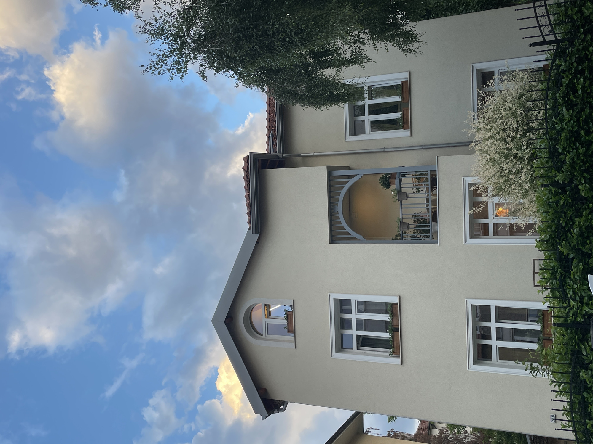 The image captures the exterior of a light-colored building, likely a house or apartment, viewed from a low angle against a partly cloudy sky. The building's facade is a neutral, possibly beige or light yellow, color, contrasting with the bright white frames of its numerous windows.
Several rectangular windows are visible, some equipped with window boxes holding green plants. One window on an upper floor features a distinctive arched top. A prominent central feature is a recessed balcony or small terrace, enclosed by a grey metal railing with an elegant arched design at the top and vertical bars. Lights are illuminated within this balcony area and in some of the visible windows, suggesting it is late afternoon or early evening. The roof, partially visible on the left, is covered with reddish-brown tiles, and a grey rain gutter runs along its edge.
Above the building and on its left, a leafy tree with dense green foliage extends into the frame. To the right of the building, a black metal fence with decorative scrolls is heavily intertwined with lush green climbing plants and a unique bush featuring variegated white and green leaves. The sky overhead is a blend of clear blue and scattered clouds, some of which are illuminated with a warm, golden light, indicative of the setting sun. The scene is static, devoid of people or direct activity, conveying a peaceful, residential atmosphere in what is stated to be Sofia, Bulgaria. No text is visible in the image.