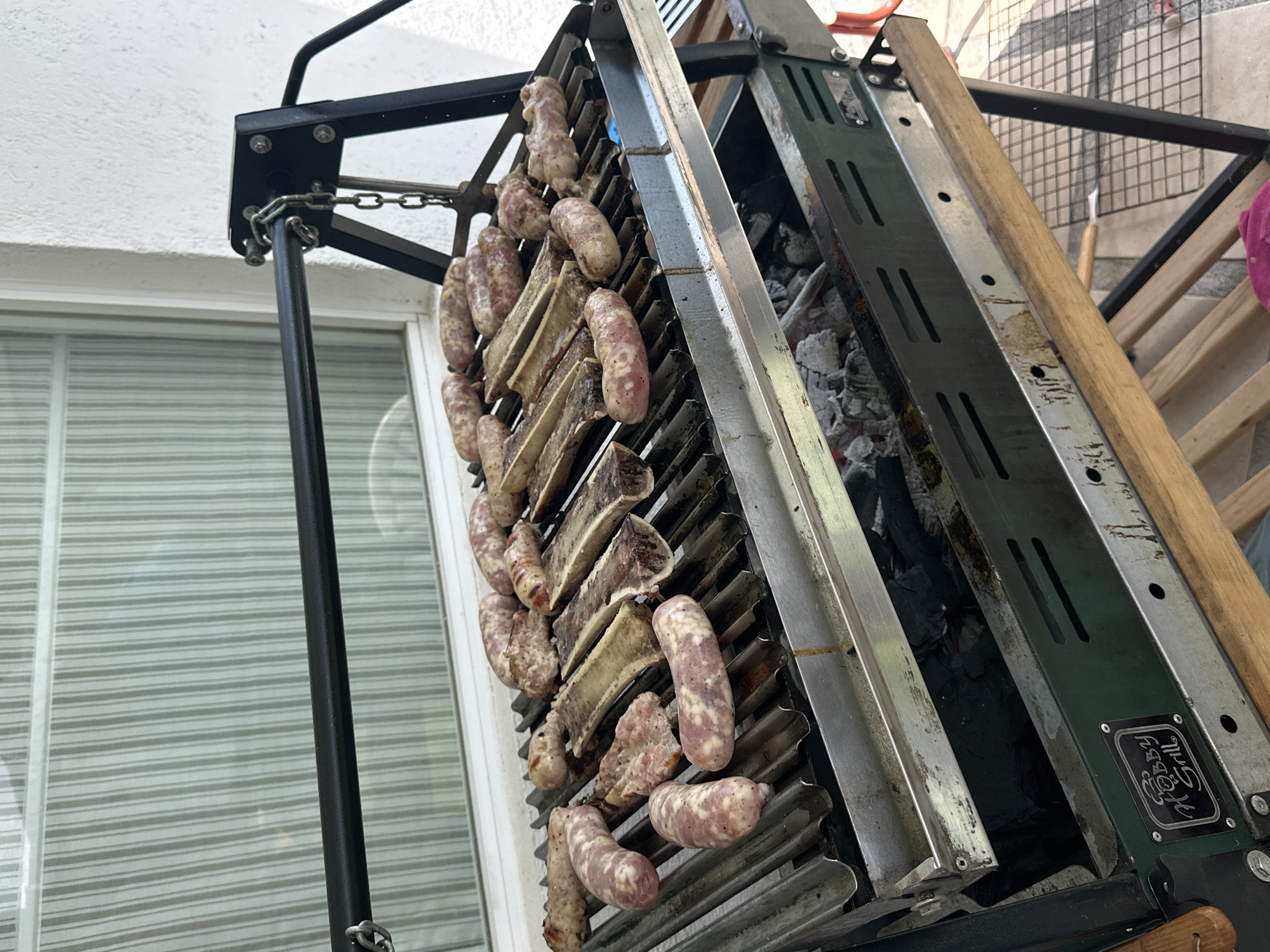 A grill loaded with sausages and ribs is seen at an angle. The grill is made of metal and has a chain attached to the top. There are hot coals in the bottom of the grill. The location is Cháhuac, Mexico.