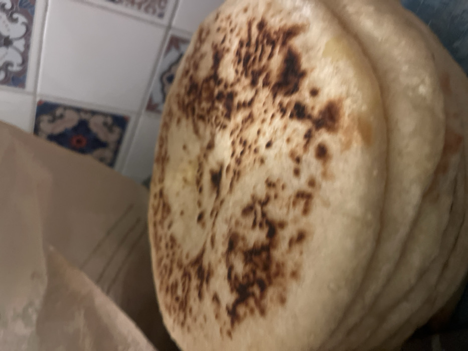 A stack of flatbreads is the focal point, featuring a lightly browned surface with darker speckles. Their soft texture is implied by the visible folds and slight overlapping of each piece. To the left, a nondescript, beige material hints at packaging or a background element. The backdrop is a section of tiled wall, adorned with decorative patterns in blue, white, and red, adding a touch of visual interest. The overall impression is simple and focused on the culinary subject matter.