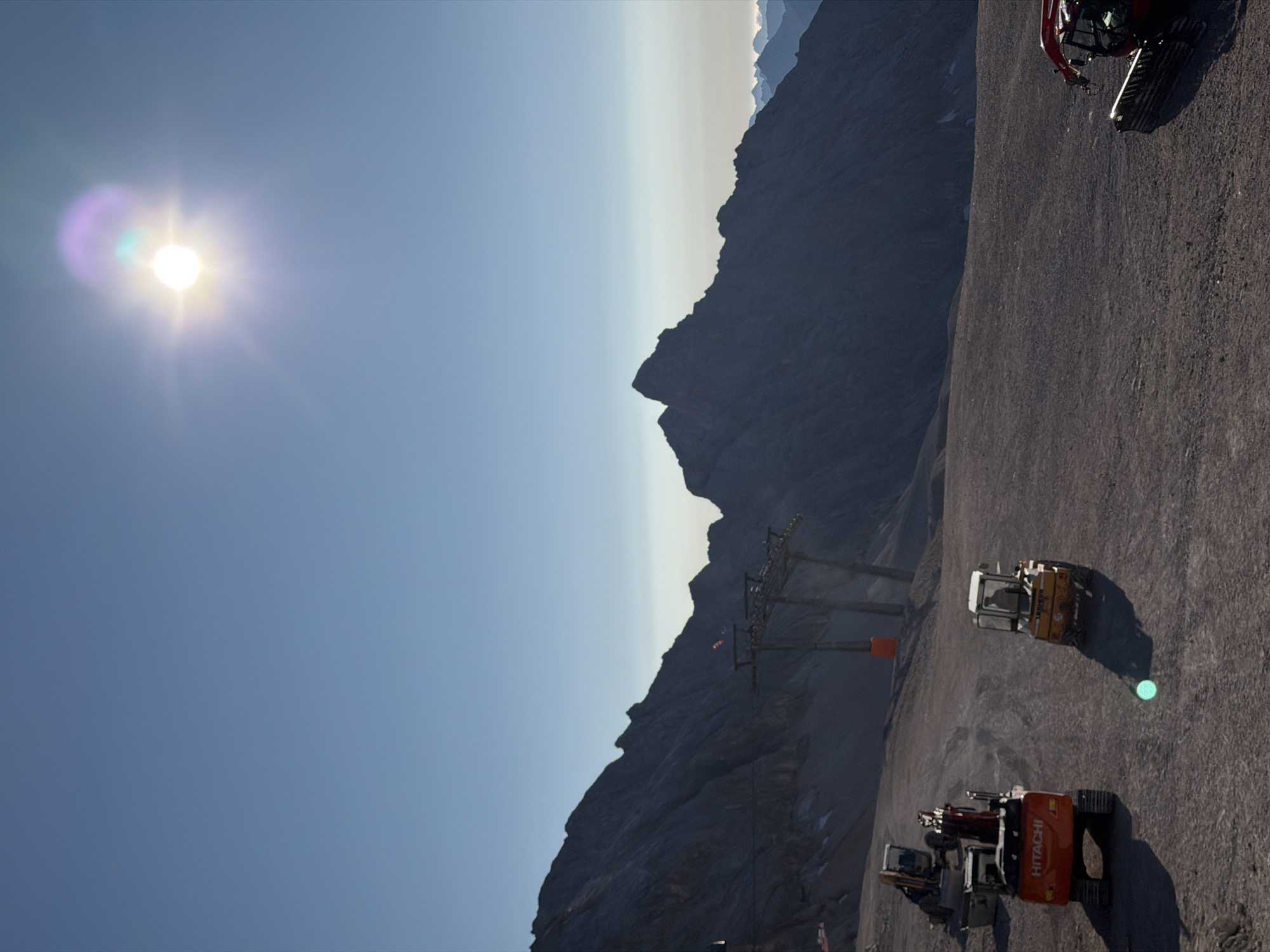 The image depicts a mountainous landscape with a clear blue sky and bright sunlight, possibly indicating a summer day. In the foreground, there are several construction vehicles, including excavators and what appears to be a snow groomer. One excavator is orange and marked with "HITACHI." Another vehicle is yellow and appears to be a small loader. The terrain is rocky and barren, suggesting a high-altitude location. In the background, there are steep, dark mountains, and the skeletal structures of a ski lift are visible, with pylons and cables. The overall scene suggests ongoing construction or maintenance work in a ski resort area, possibly during the off-season. The presence of ski lift infrastructure strongly points to a location within or near the Zugspitze massif, the highest peak in Germany, which is located near Garmisch-Partenkirchen.