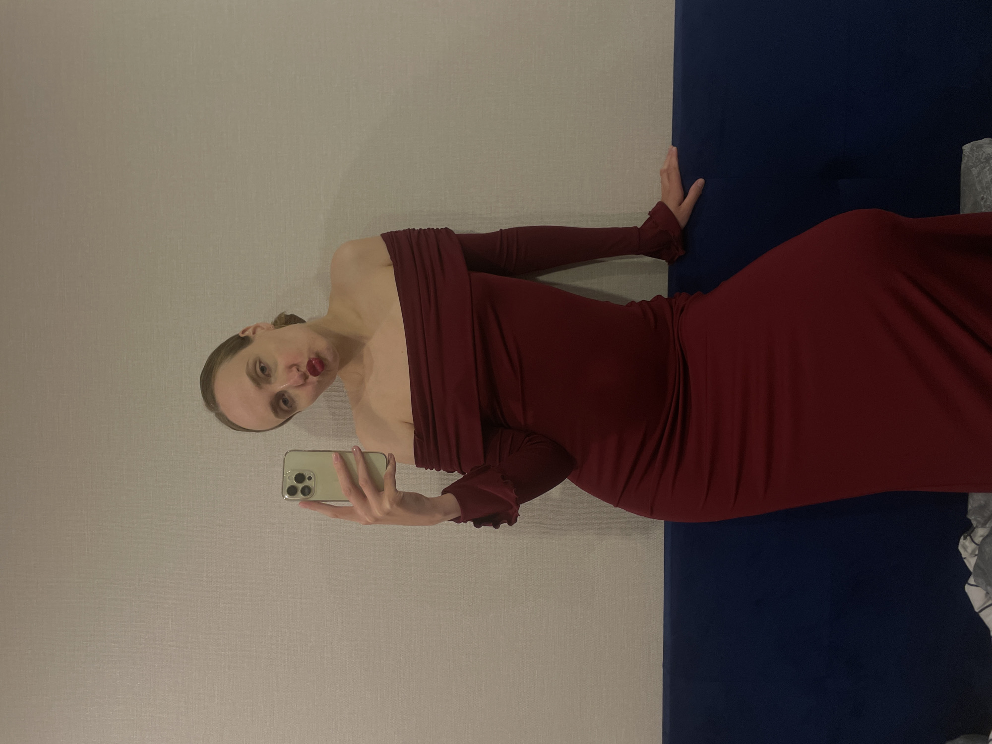 A woman in a dark red dress is taking a selfie. She is holding a phone with her left hand. She is standing against a wall that is light gray on the left and dark blue on the right. Her right hand is touching the dark blue wall. She is wearing dark red lipstick. The location is undetermined.